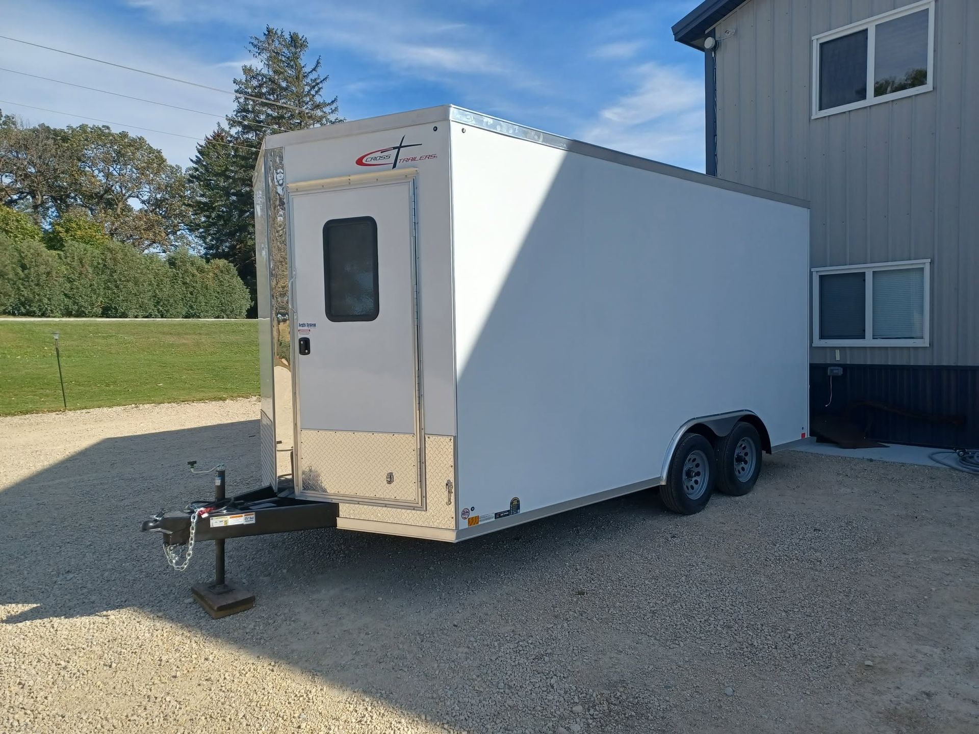 White enclosed trailer with double axles parked beside a gray building on a gravel lot.