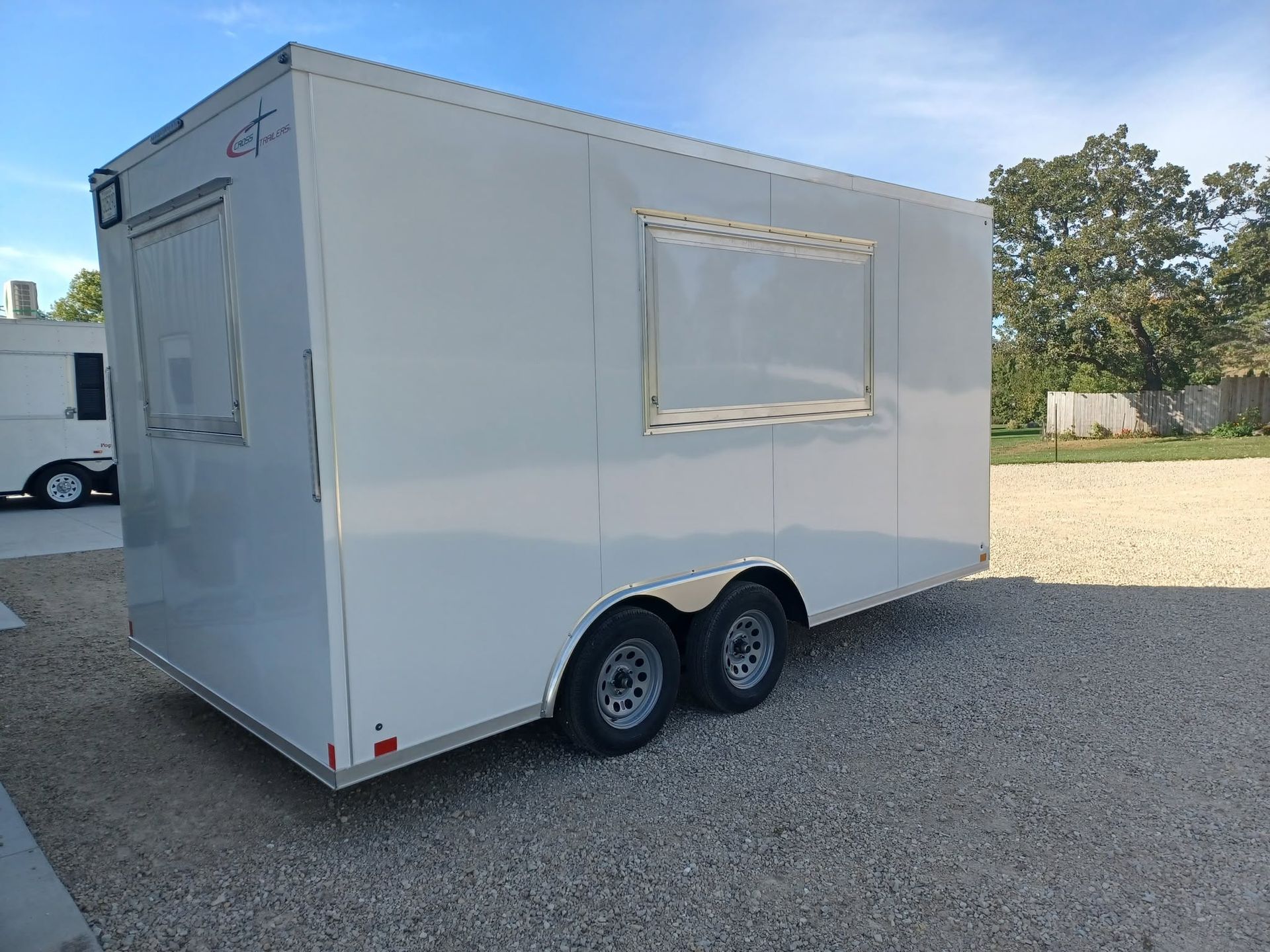 White dual-axle enclosed trailer with side windows parked on gravel under a blue sky.