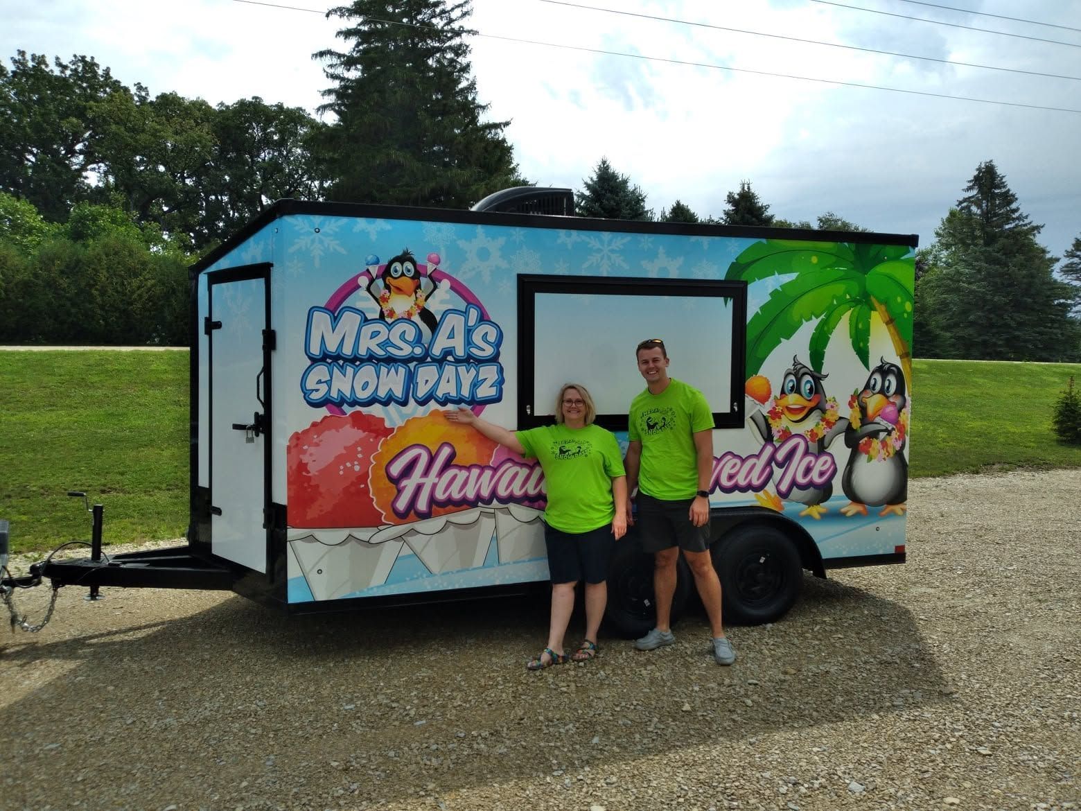 Two people standing by a colorful shaved ice trailer in a grassy outdoor setting