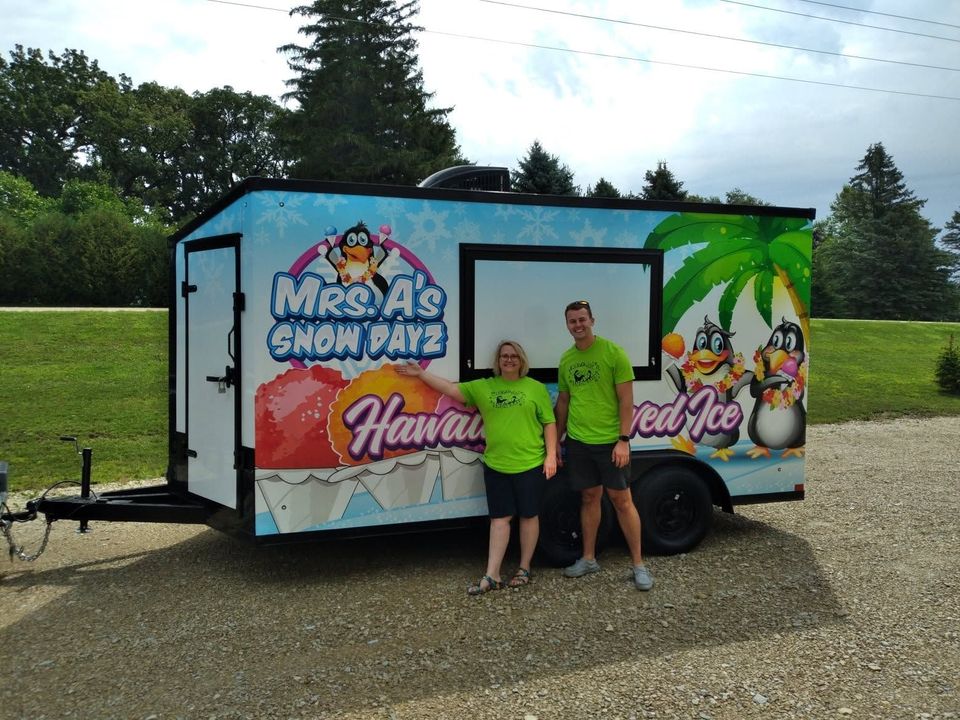 Two people stand beside a colorful shaved ice trailer on a gravel lot.