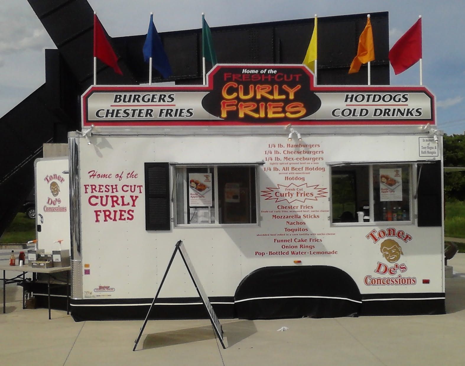 White food stand with black awning advertising curly fries, burgers, and hot dogs on a sidewalk.