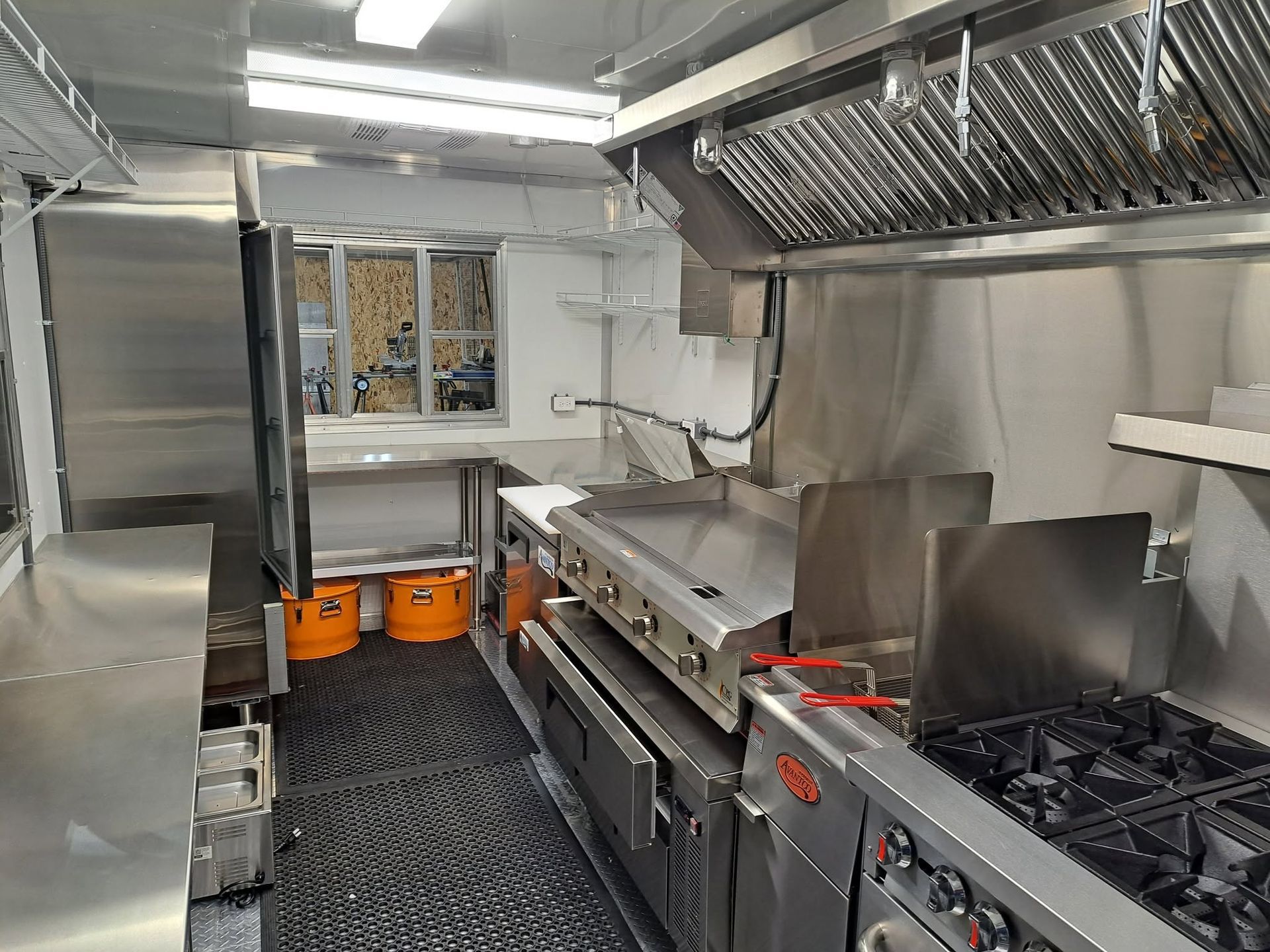 Stainless-steel commercial kitchen with stovetops, prep counter, ovens, and orange buckets in the back.