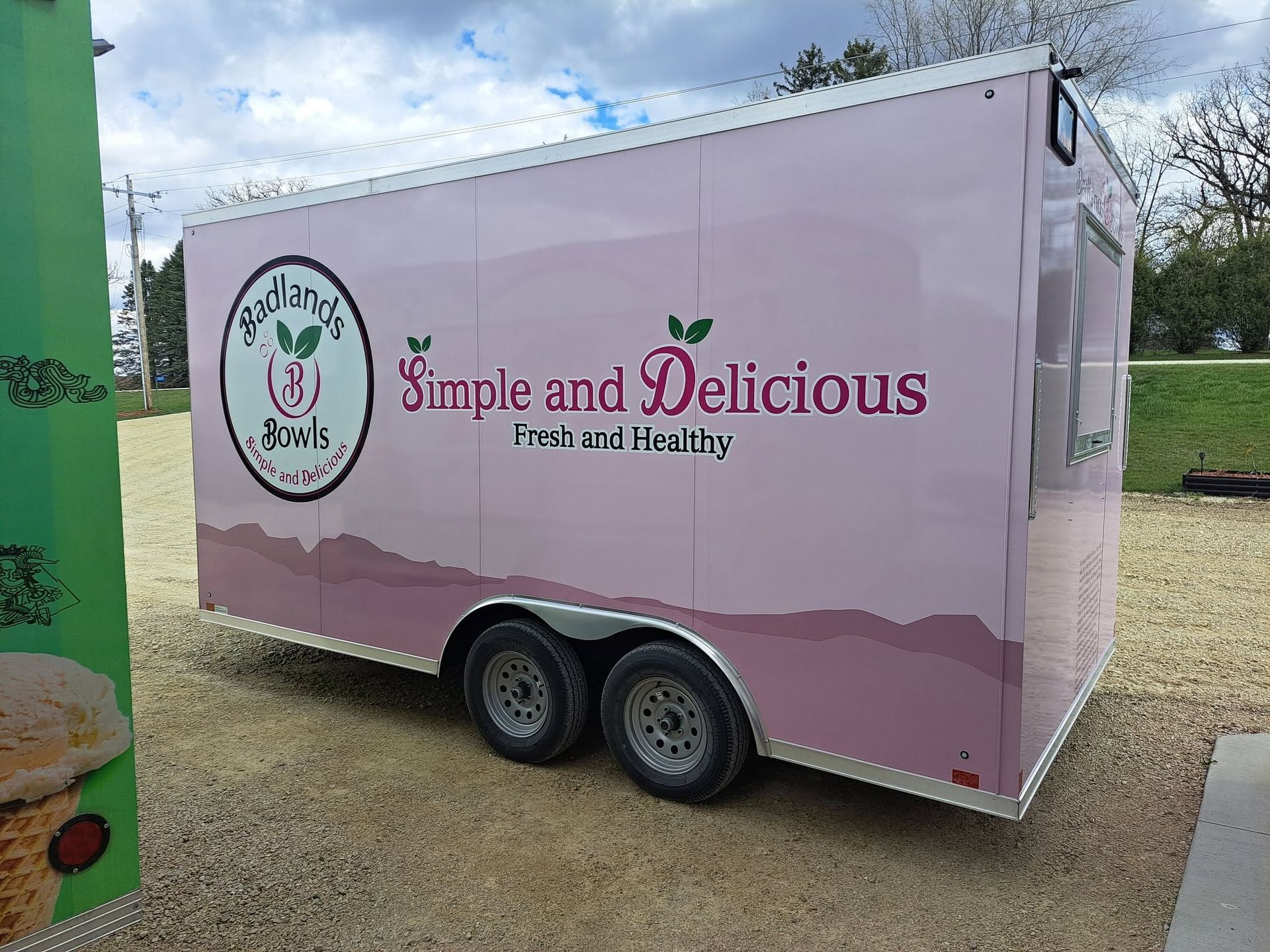 Pink food trailer with “Simple and Delicious” branding parked on gravel outdoors