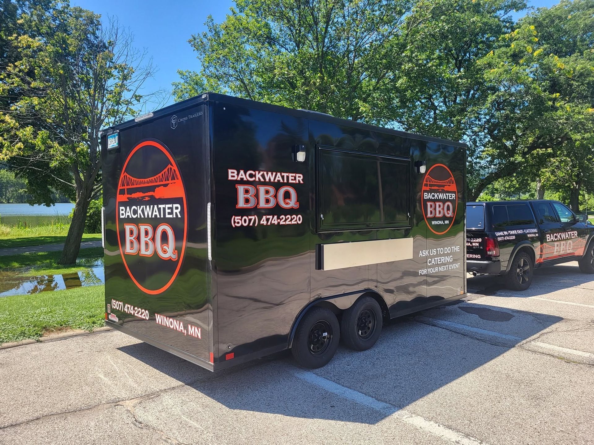 Black BBQ food trailer with red logos parked outdoors beside a vehicle and trees