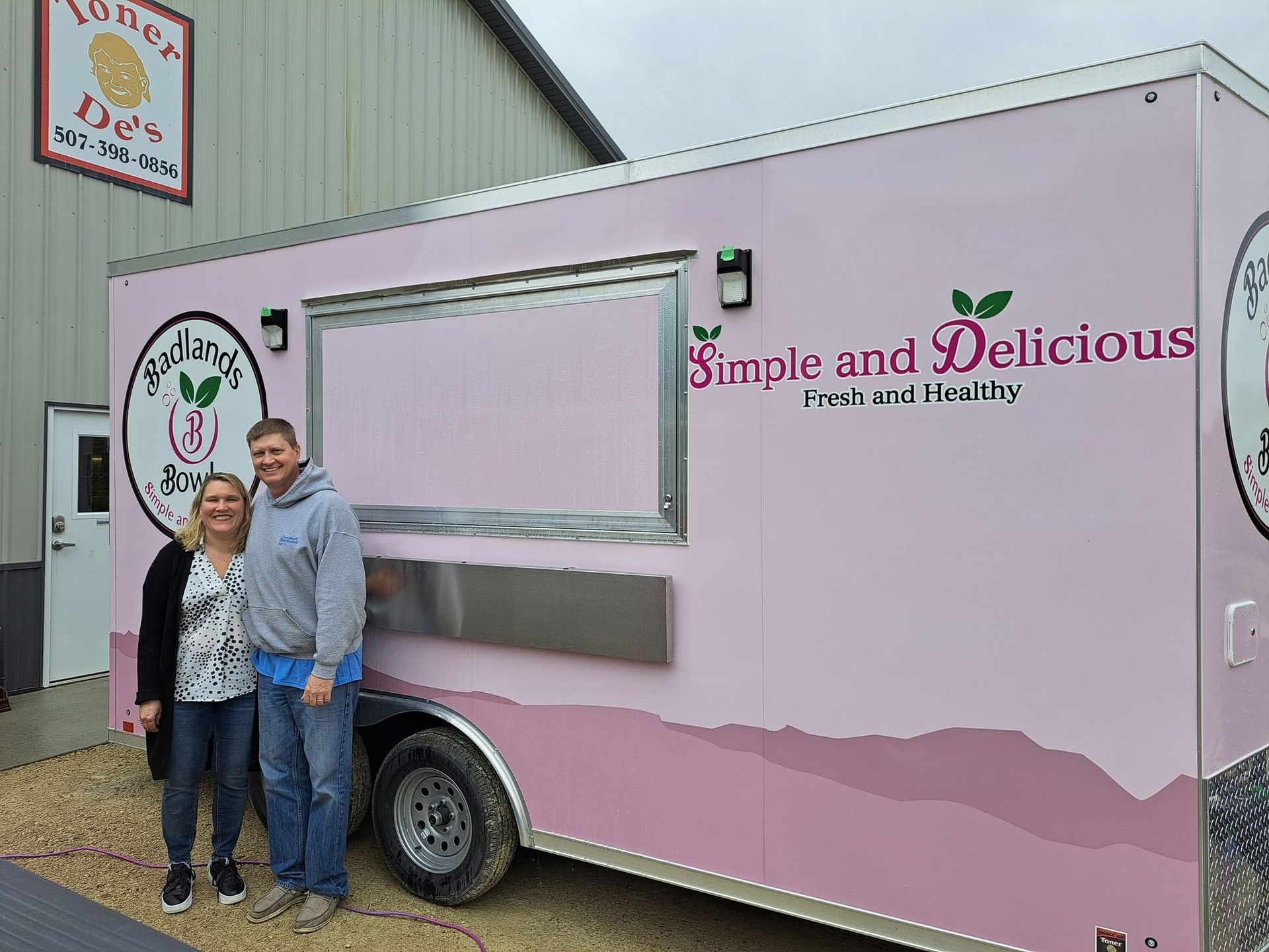 Two people standing beside a pink food trailer labeled “Simple and Delicious”