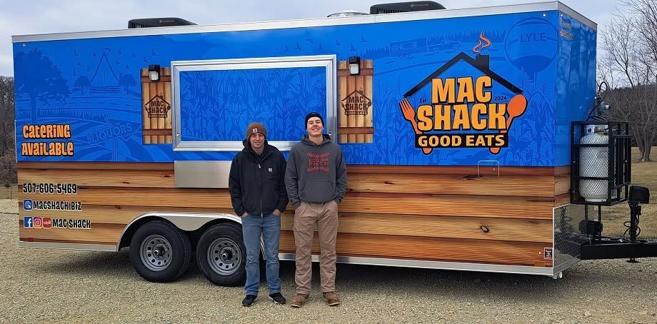 Two people stand beside a blue-and-wood Mac Shack Good Eats food trailer