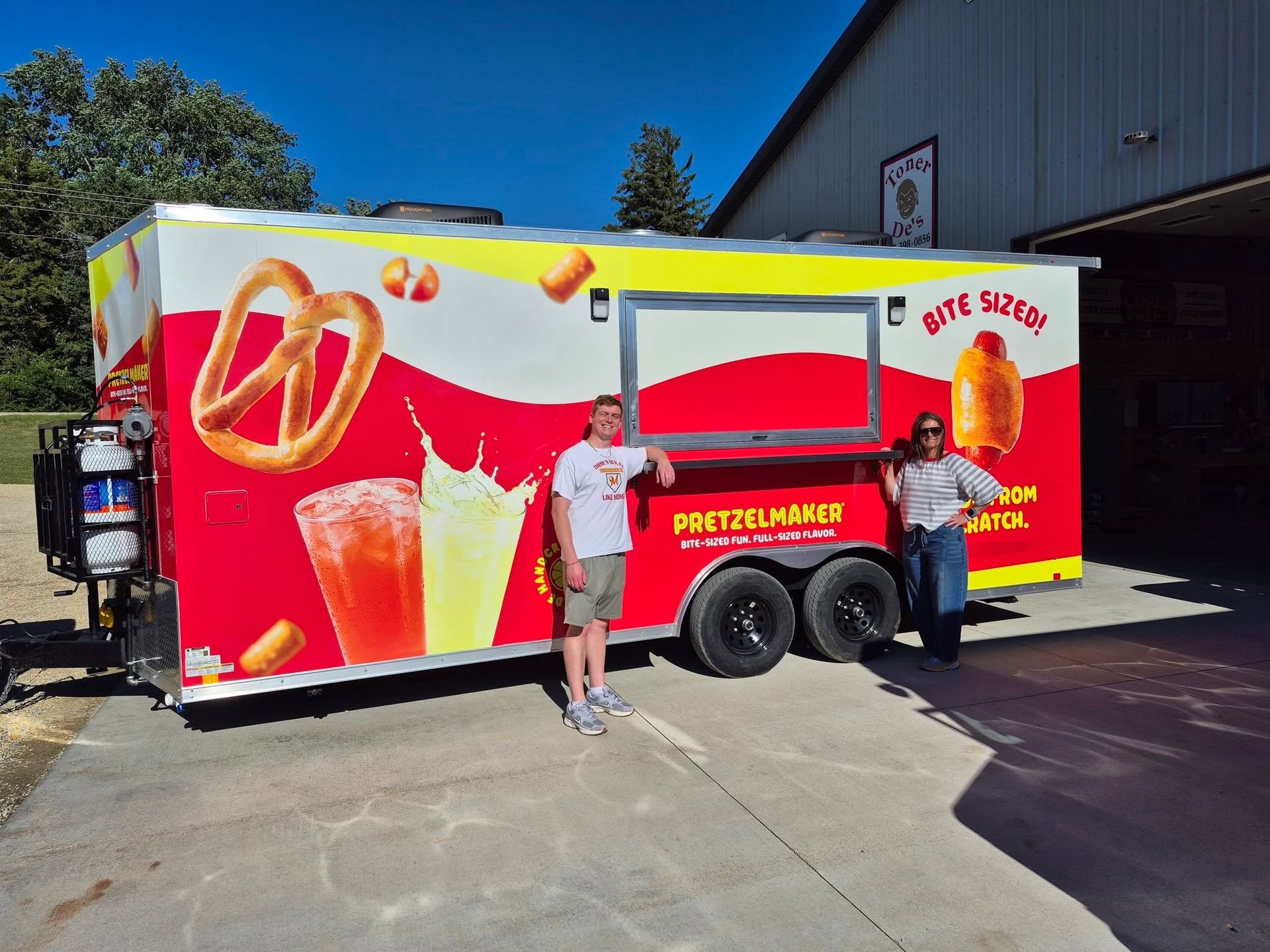 Two people standing by a red food trailer with pretzel graphics in a sunny parking lot