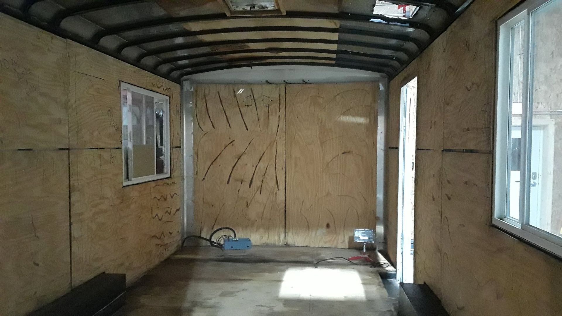 Empty unfinished room with plywood walls, exposed beams, and windows letting in daylight.