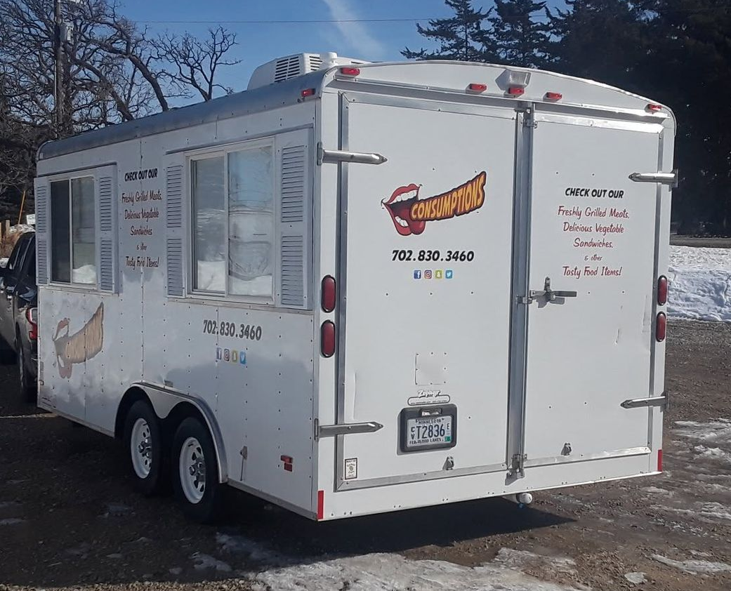 White food truck trailer with menu signs, parked outdoors in a wooded area.
