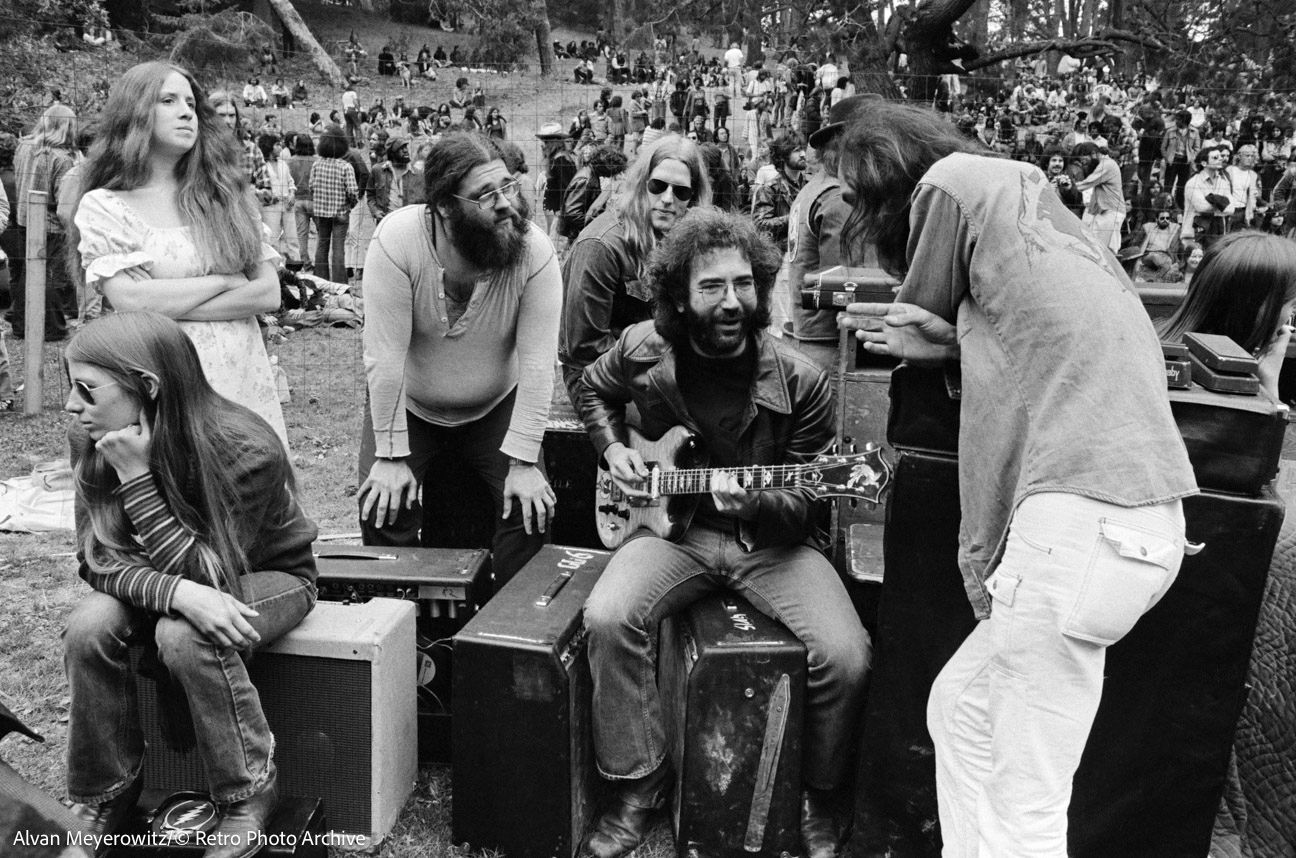 Musicians sitting and standing outdoors at a crowded festival, playing guitar and talking together