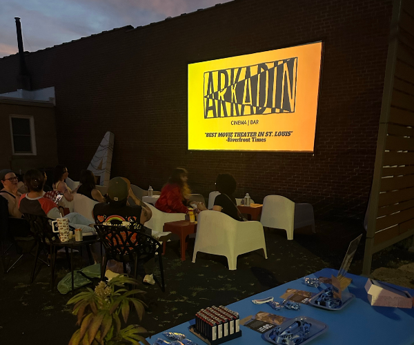 Outdoor evening screening with seated audience facing a projected yellow title slide on a brick wall.
