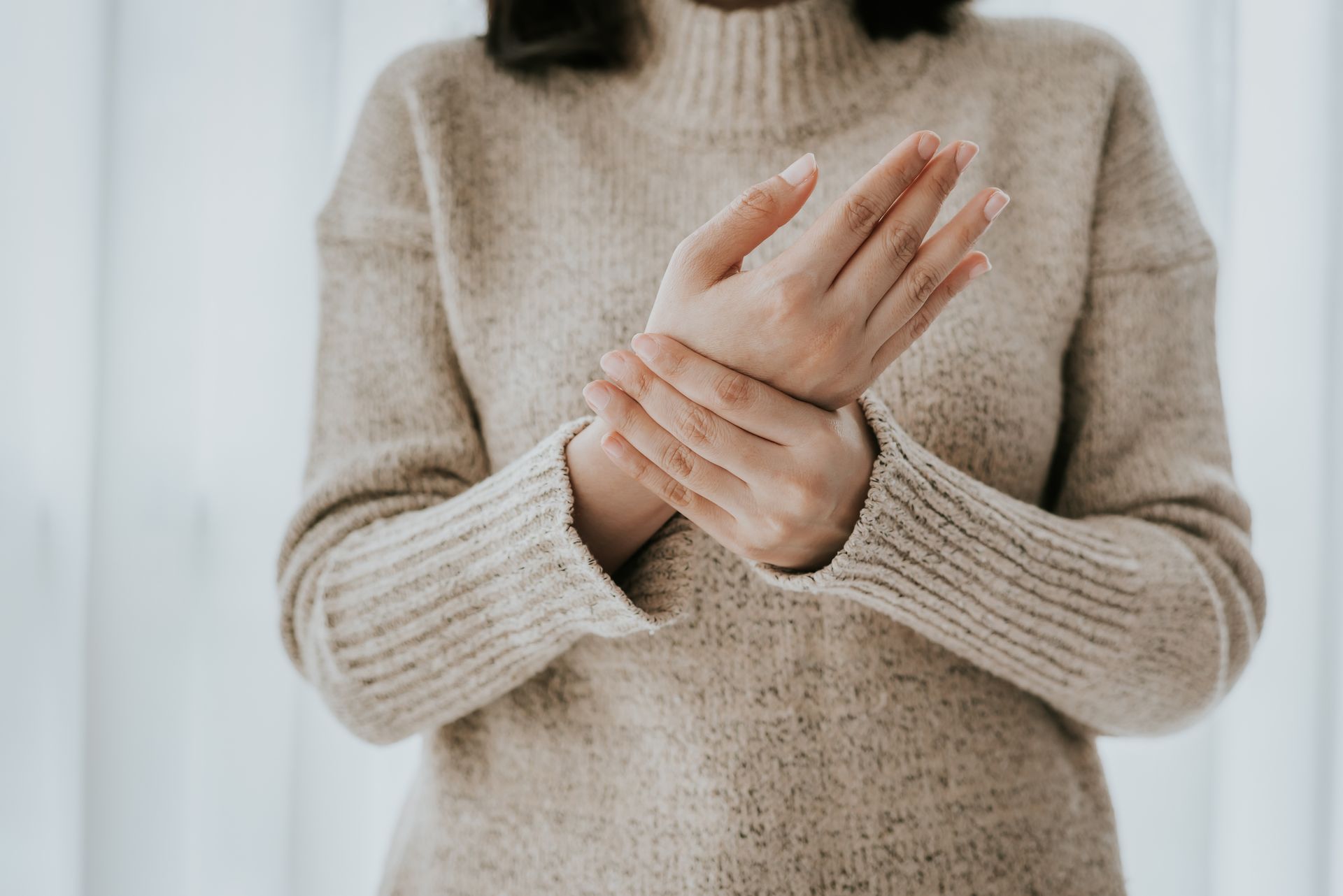 Person in a beige sweater holding and rubbing a wrist, suggesting discomfort or pain