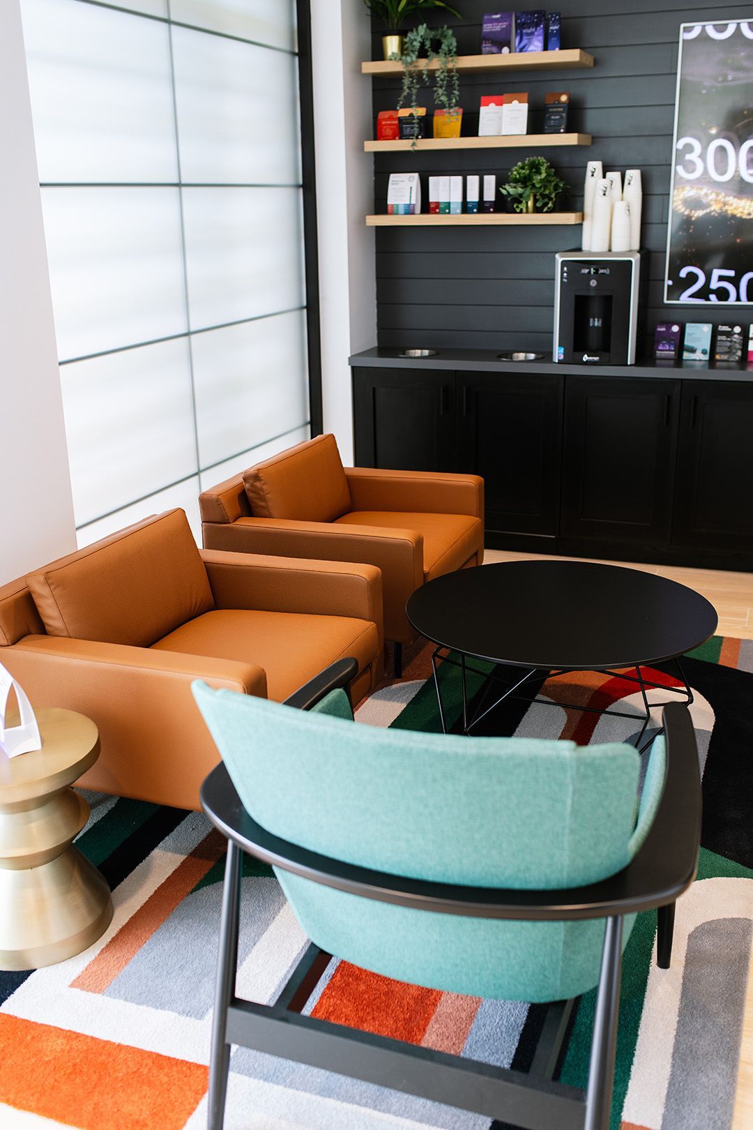 Modern lounge with teal and orange chairs, round table, and black shelving against a windowed wall