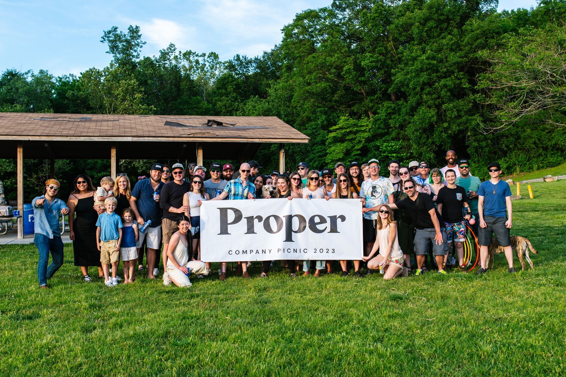 Group of people outdoors holding a “Proper” banner in front of a pavilion on a grassy field