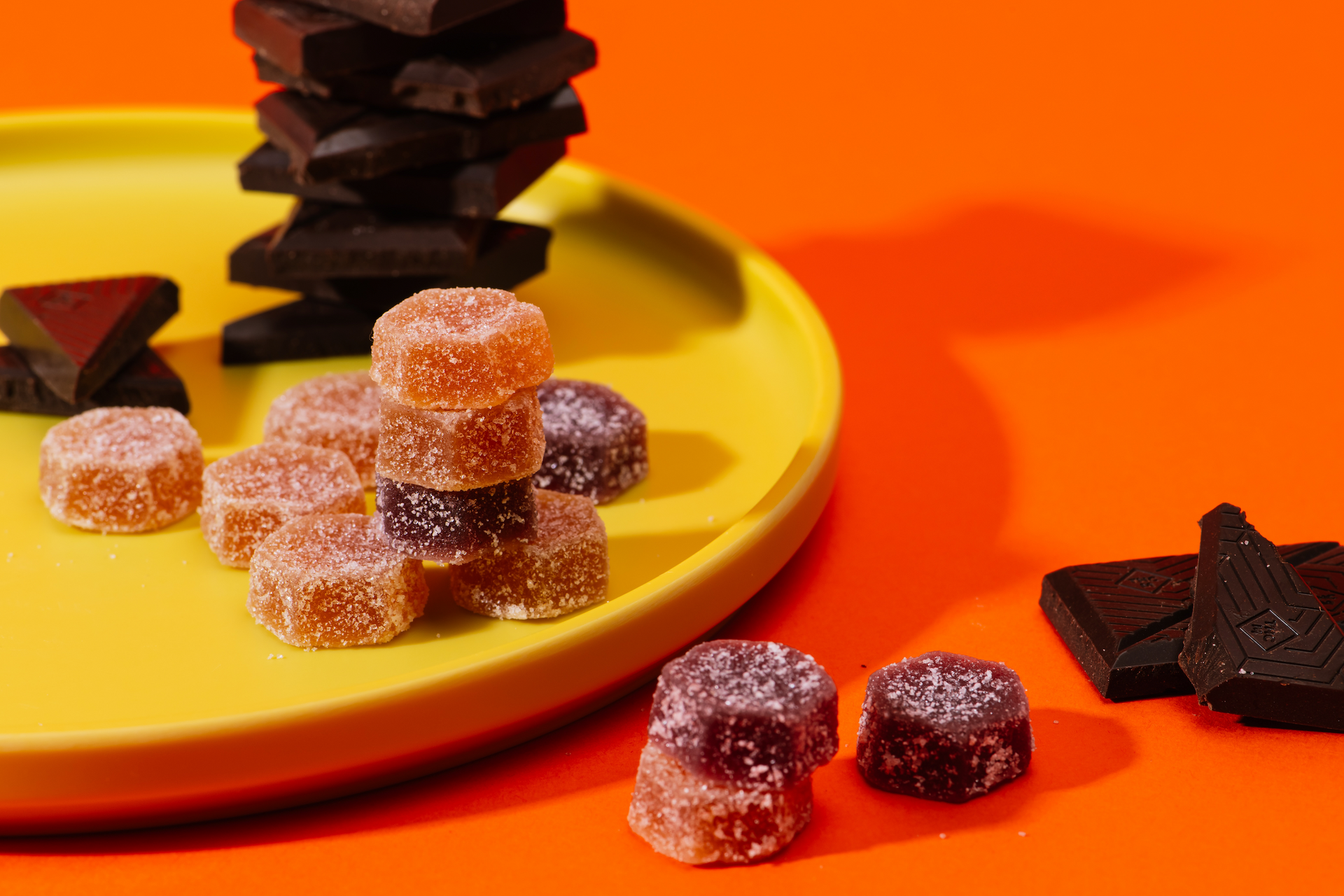 Assorted sugar-coated gummy cubes on a yellow plate and orange surface with stacked dark chocolate pieces