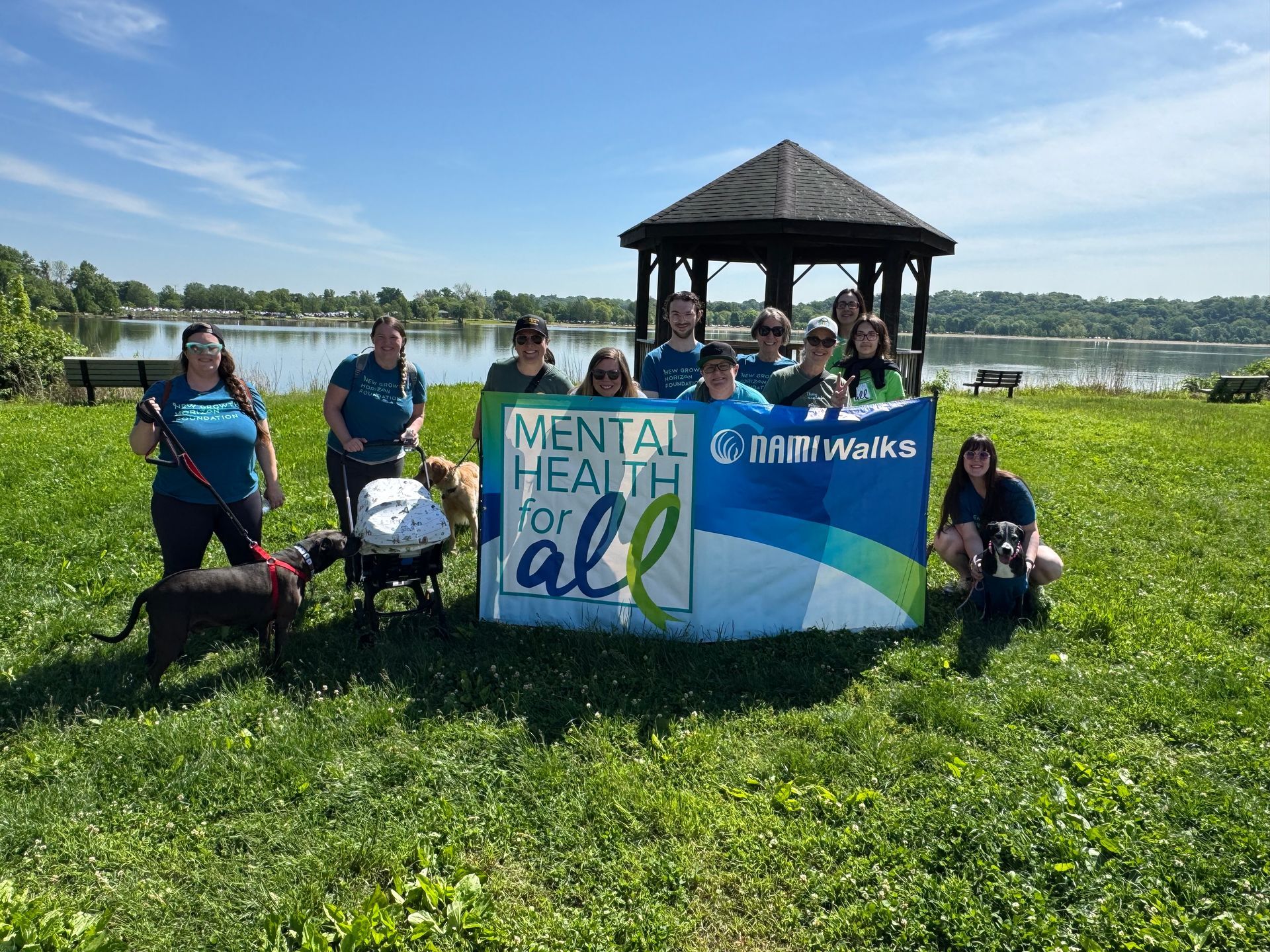 Group holding Mental Health Walk signs in a sunny park by the water with a gazebo