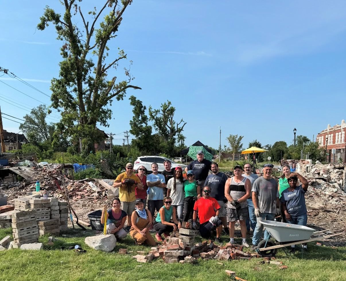 Group of volunteers cleaning debris in a grassy area under a blue sky, with rubble piles and tools nearby