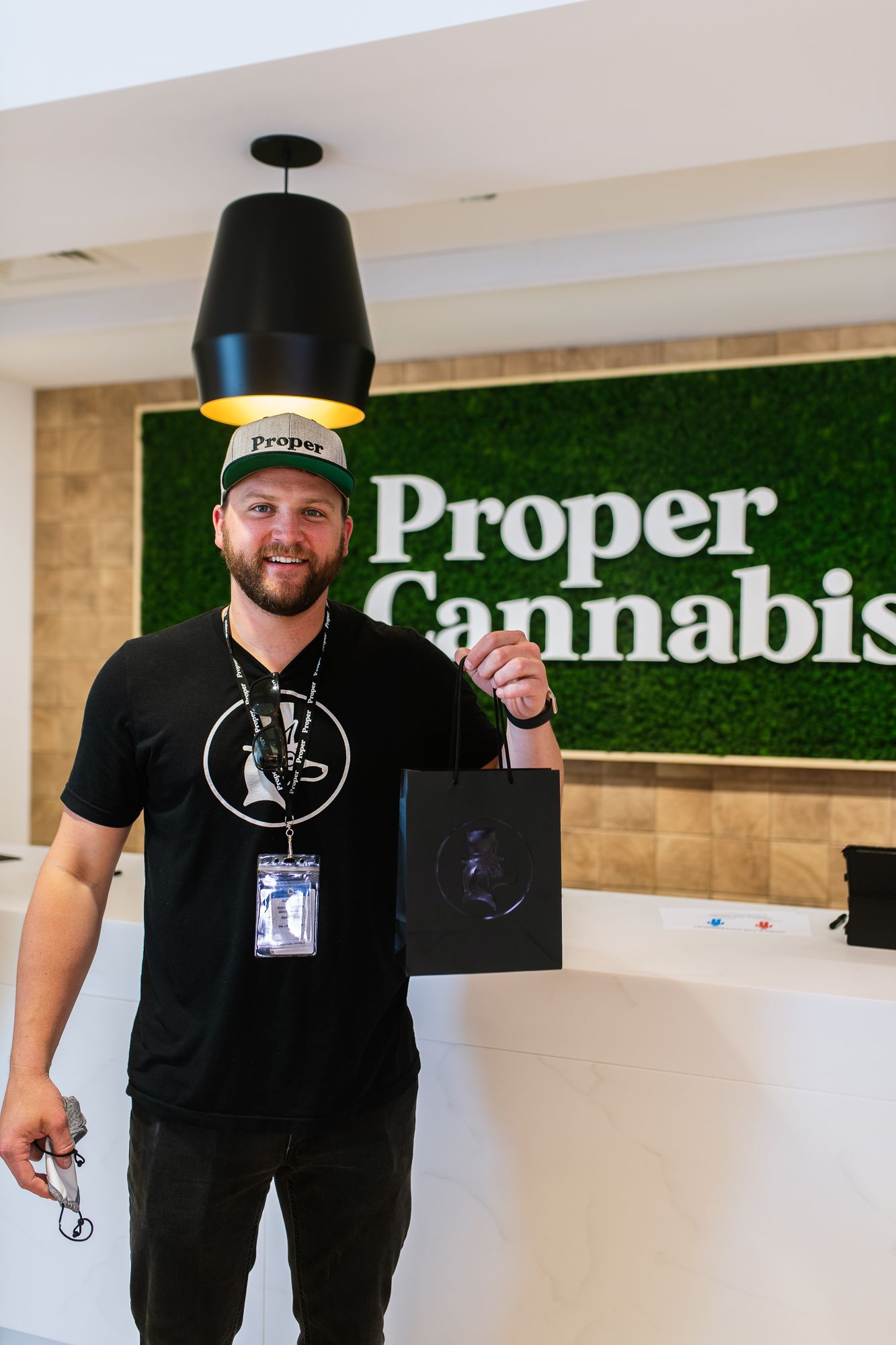 Man holding a black tote bag in front of a green “Proper Cannabis” wall display