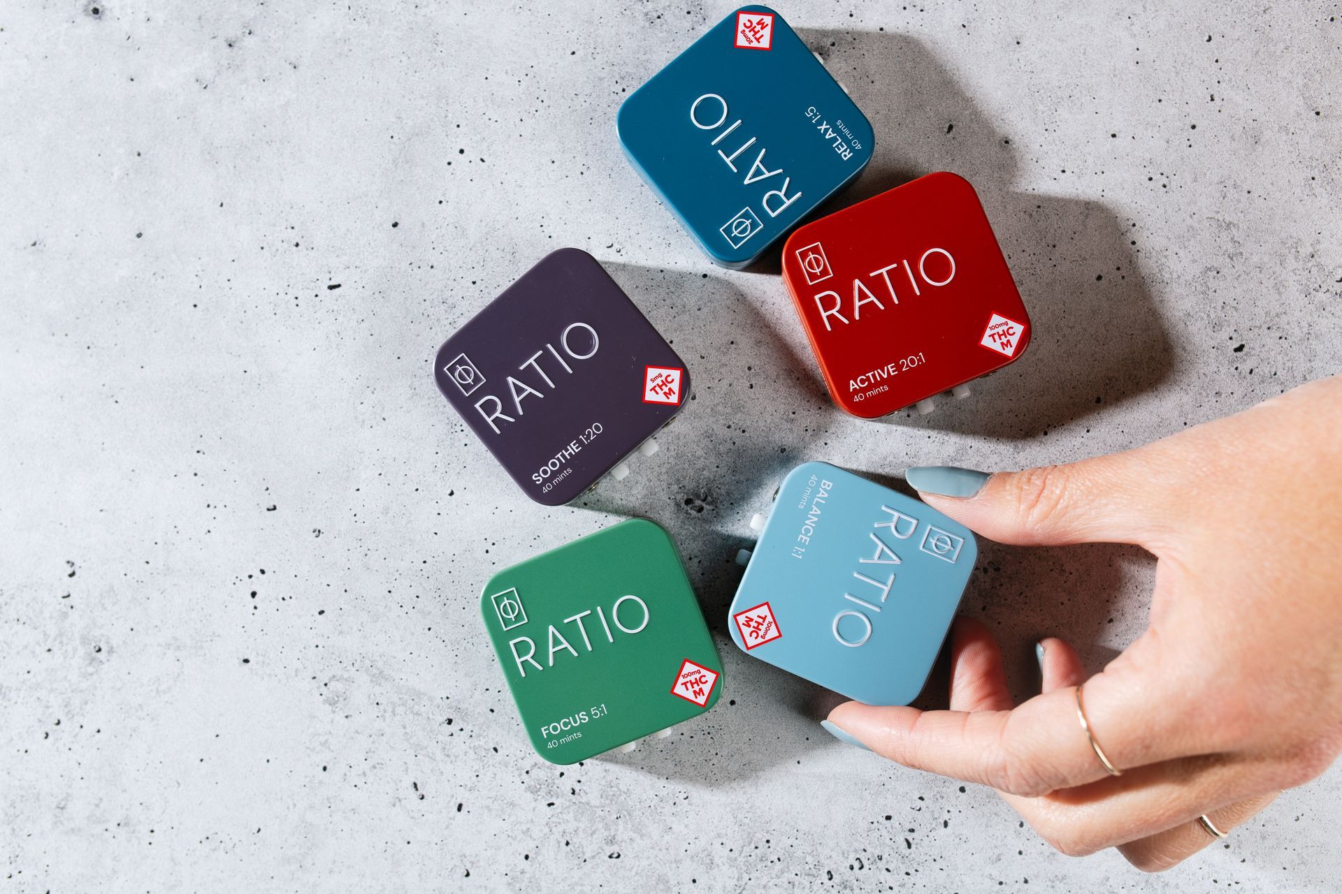 Hand holding a blue RATIO box among four colorful RATIO boxes on a concrete surface