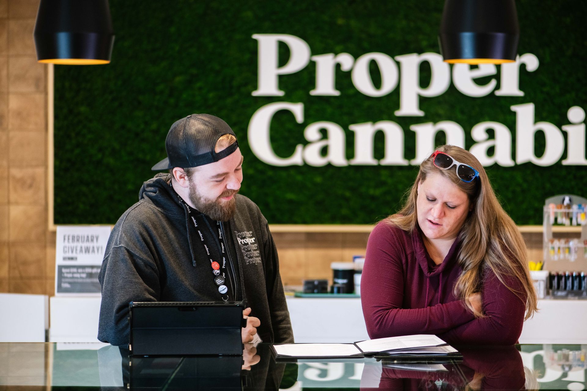 Two people talking at a cannabis shop counter beneath a “Proper Cannabis” sign.