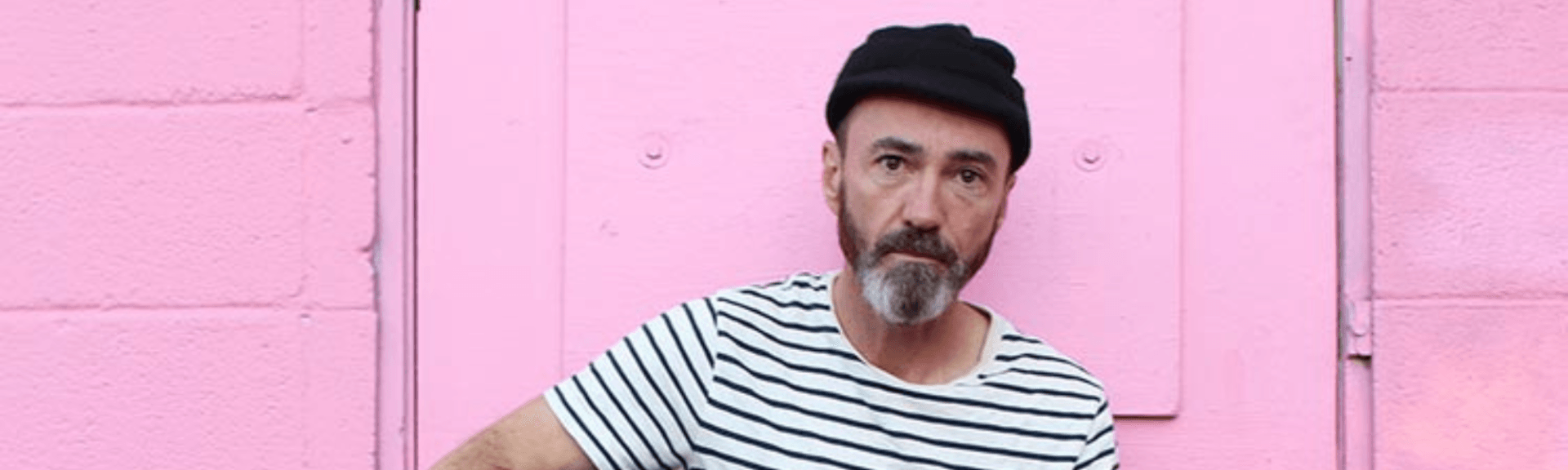 Man in black cap and striped shirt standing before a pink wall, looking at the camera