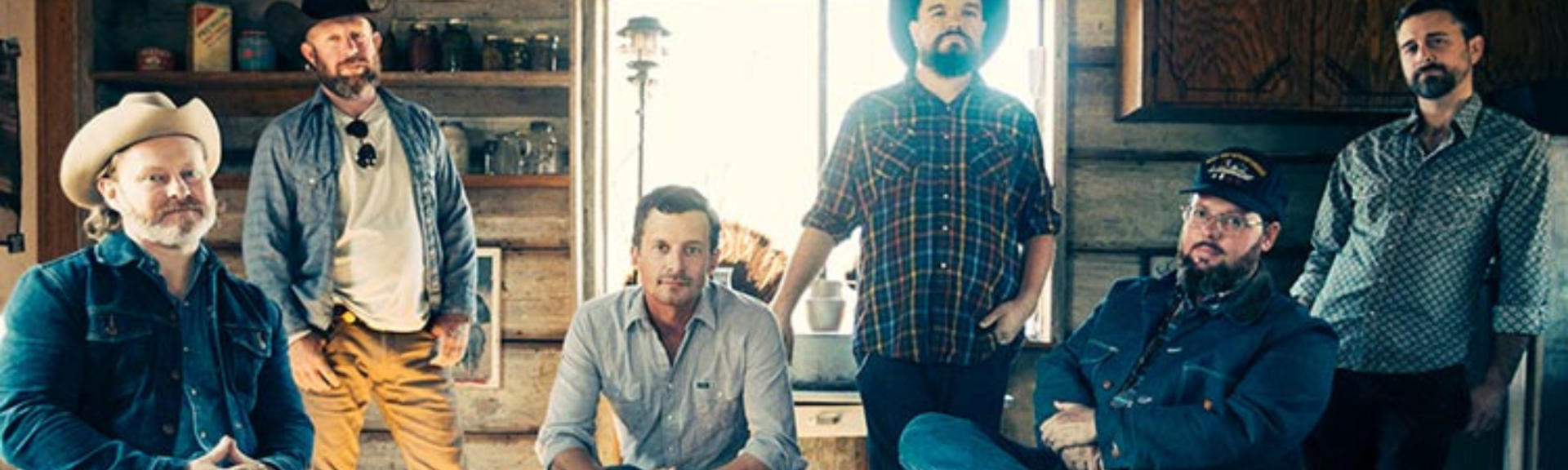 Six adults posing indoors in a warmly lit room, standing and sitting around furniture.