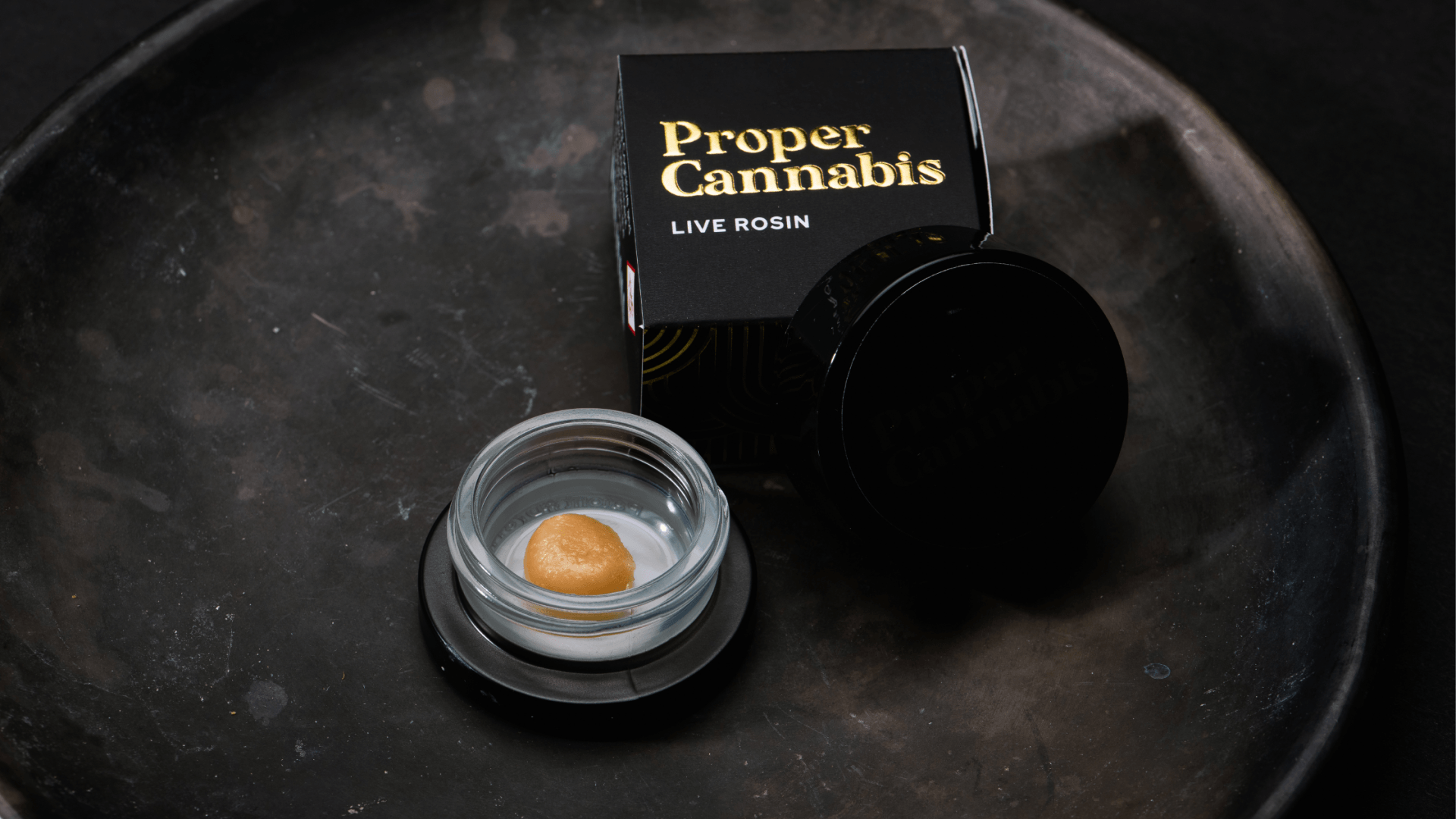 Proper Cannabis lip balm in open black tin on a dark tray, with box and cap beside it.
