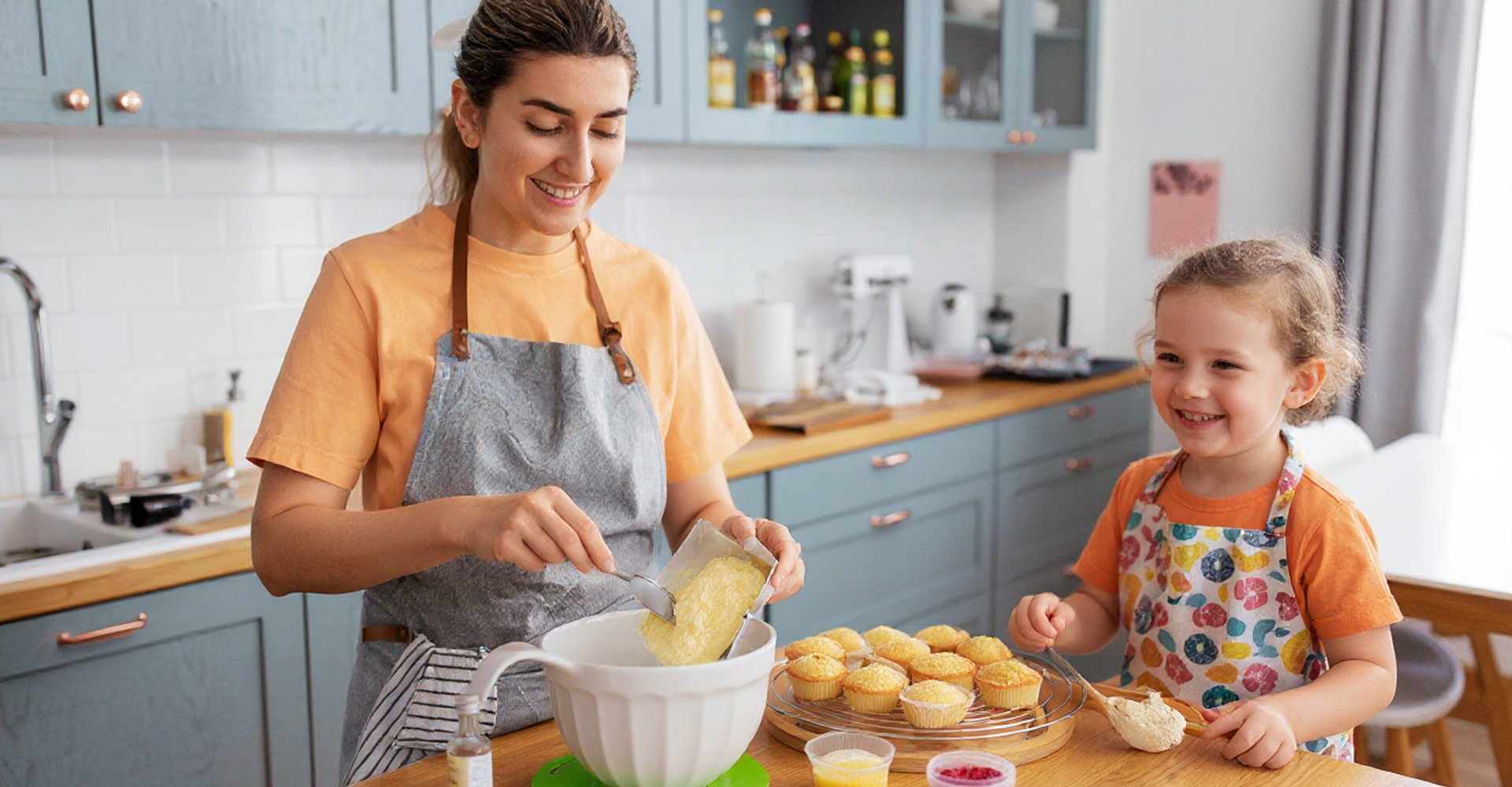 Woman and child baking in a kitchen, mixing ingredients with smiles; muffins sit on the table.