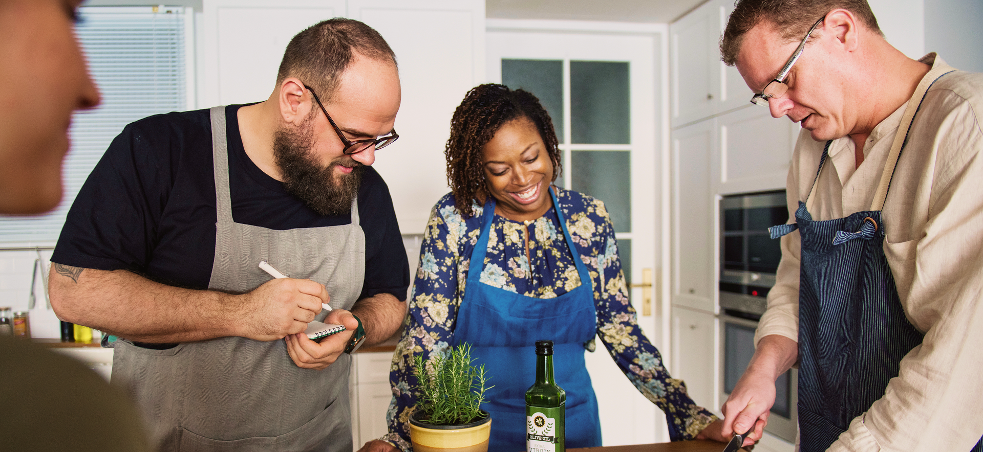 People cook together in a kitchen, wearing aprons. One man writes, another slices, a woman smiles.