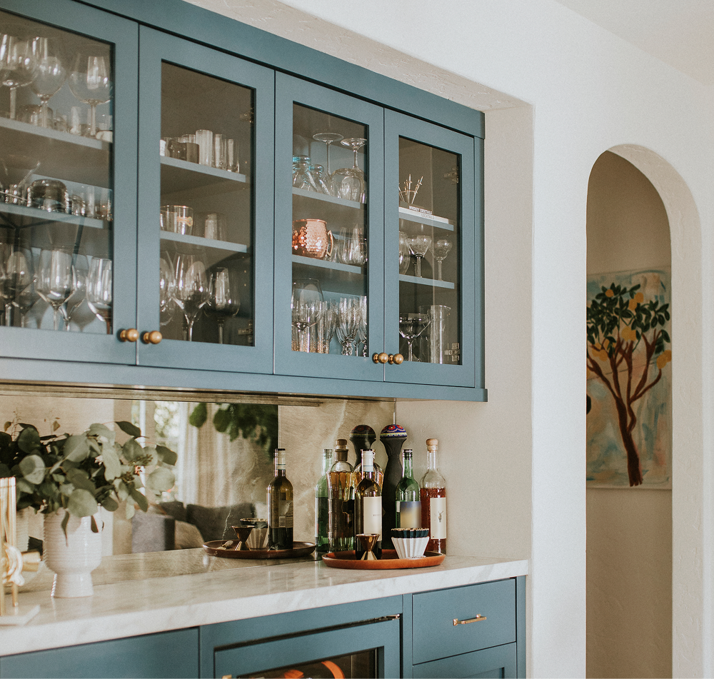 Blue bar cabinet with glass-fronted upper cabinets, marble countertop, and a mirrored backsplash.