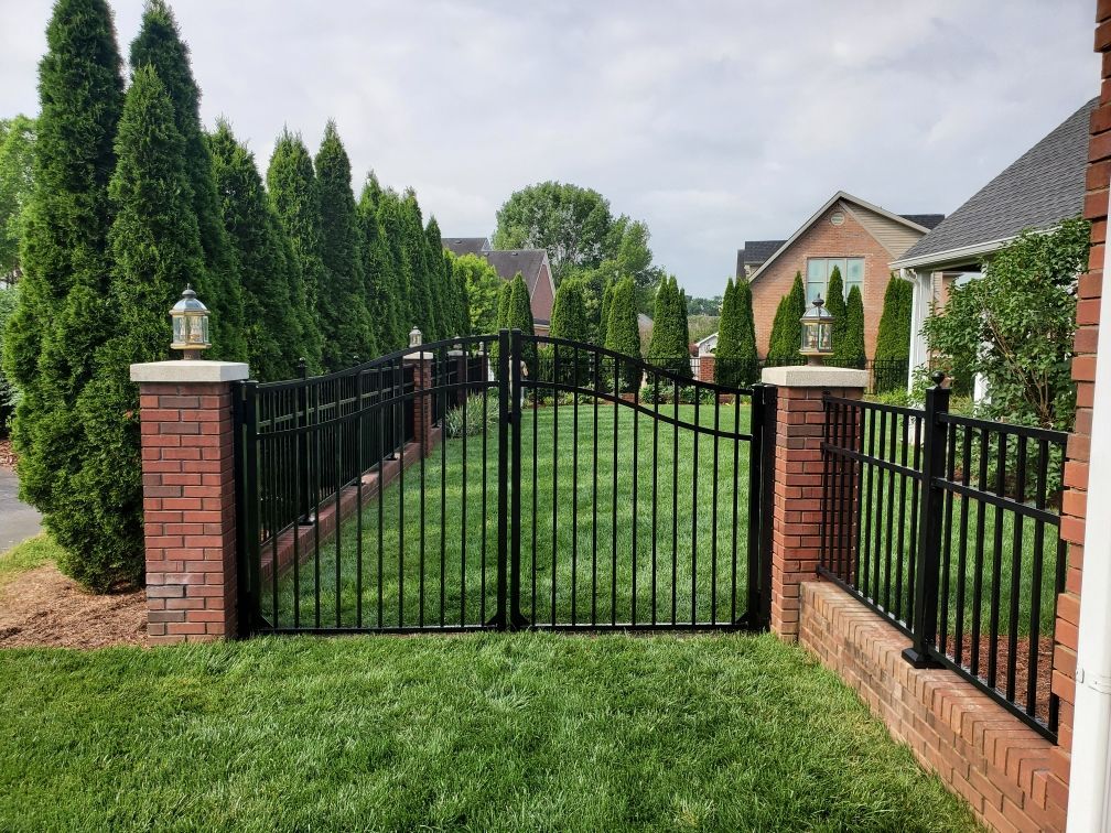 Black iron gate with brick pillars, set into a brick fence, with green grass and tall green trees.