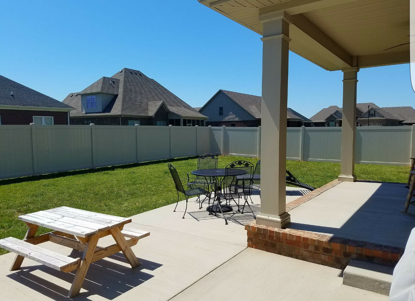 A backyard patio with a picnic table and a table with chairs, green lawn, and beige fence.