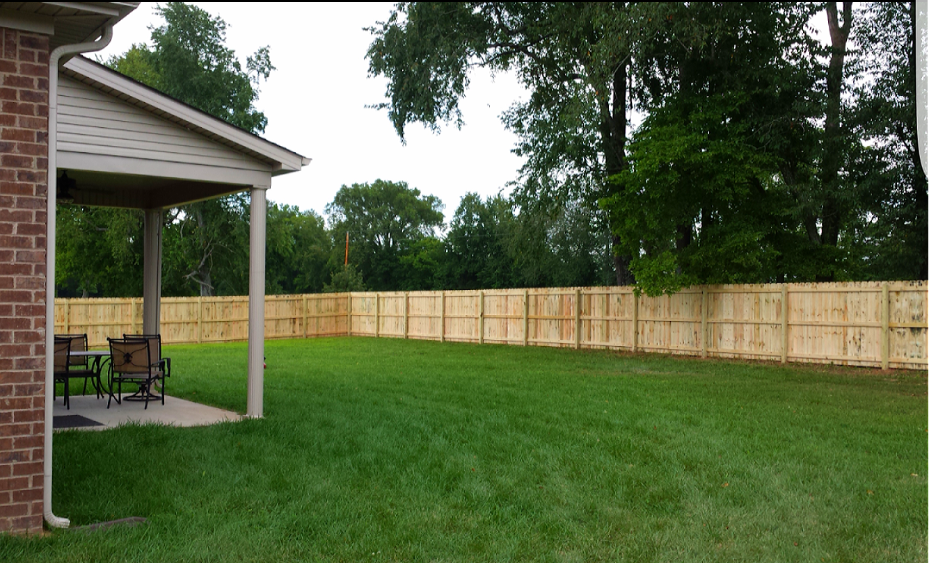 A backyard with a new wooden fence, green lawn, and a covered patio area beside a brick house.