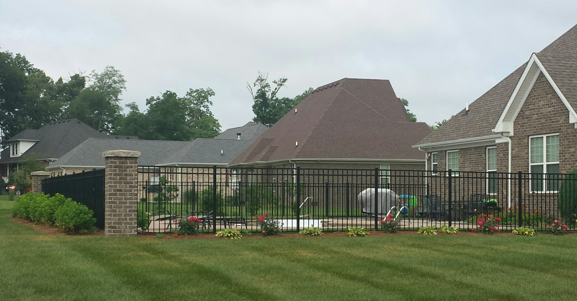 Backyard with black fence, pool, and houses under a cloudy sky. Green grass and shrubbery.