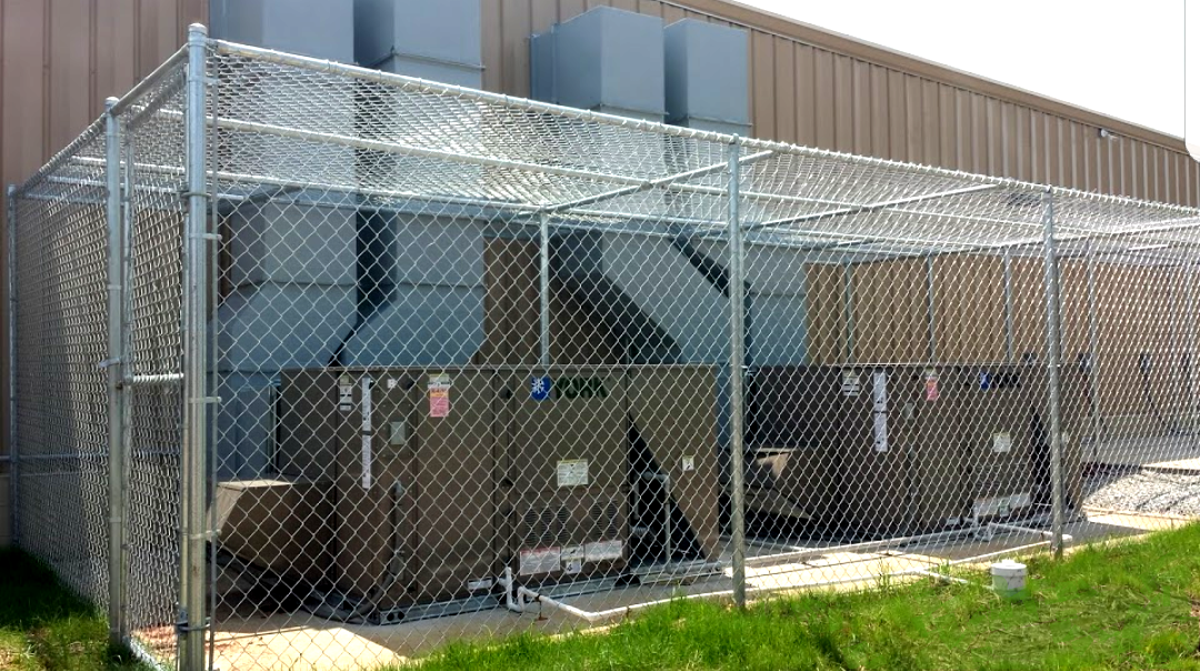 Chain-link fence encloses HVAC units outside a building with metal siding and a grassy area.