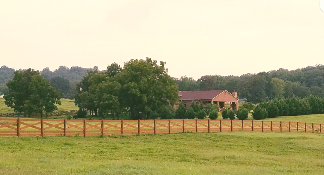 A house with a brown roof and a wooden fence in a grassy field, trees in the background.