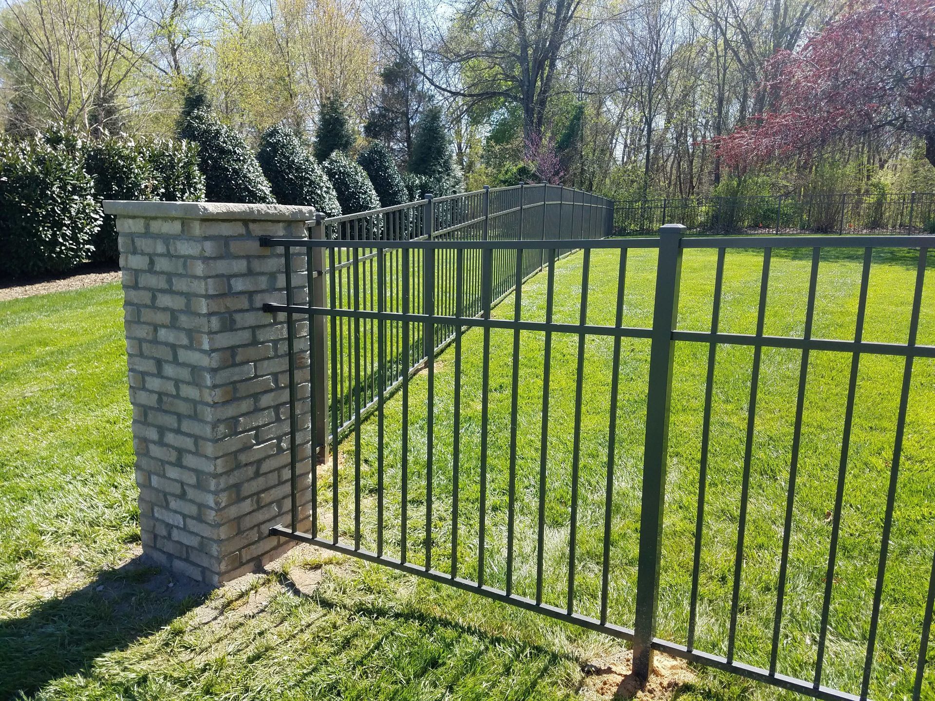 Green metal fence with brick pillar, on grass. Trees in the background, sunny outdoors.