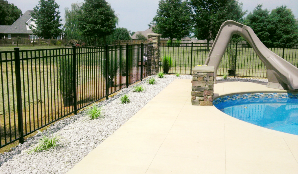 Black fence, pool with slide, tan patio, white rocks, and green plants along the fence.
