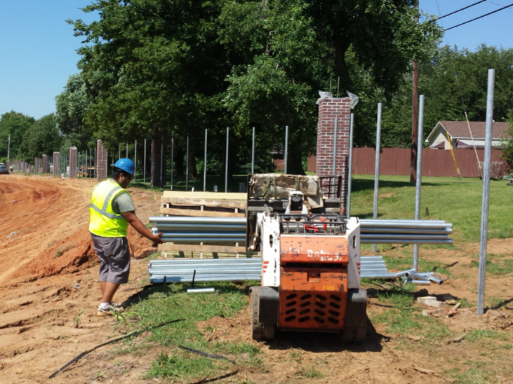 Construction worker and Bobcat constructing a fence. Worker in safety vest loads metal beams in sunny outdoor setting.