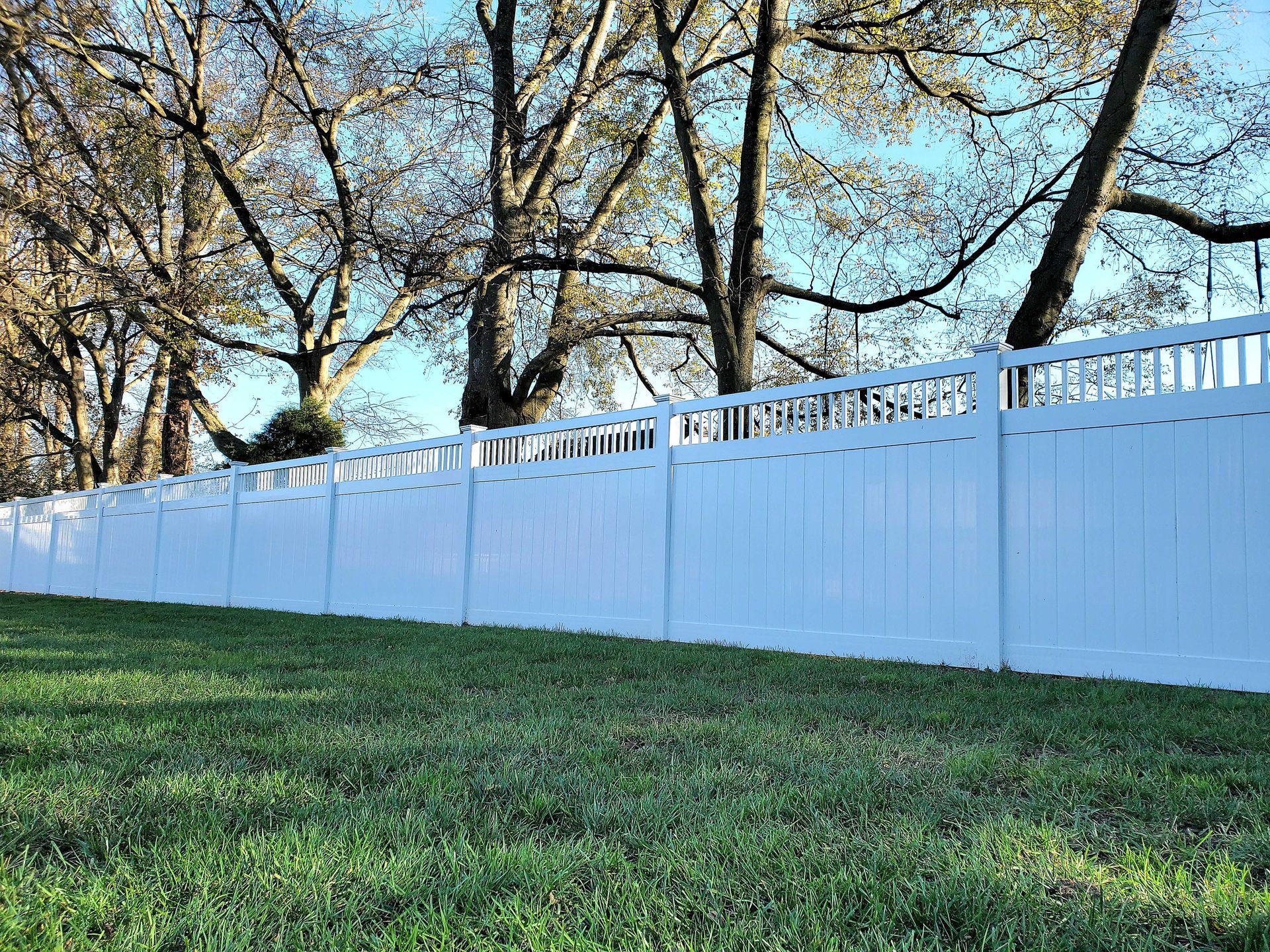 White vinyl fence on a grassy hill, with bare trees and blue sky in the background.