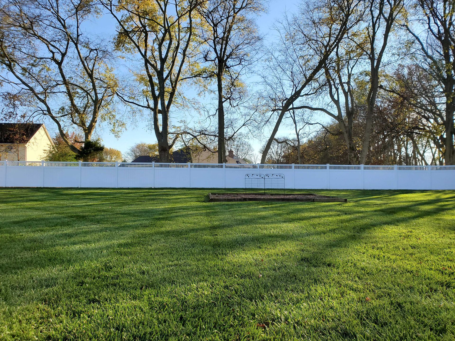 Green lawn with white fence, trees, and a house on a sunny day.