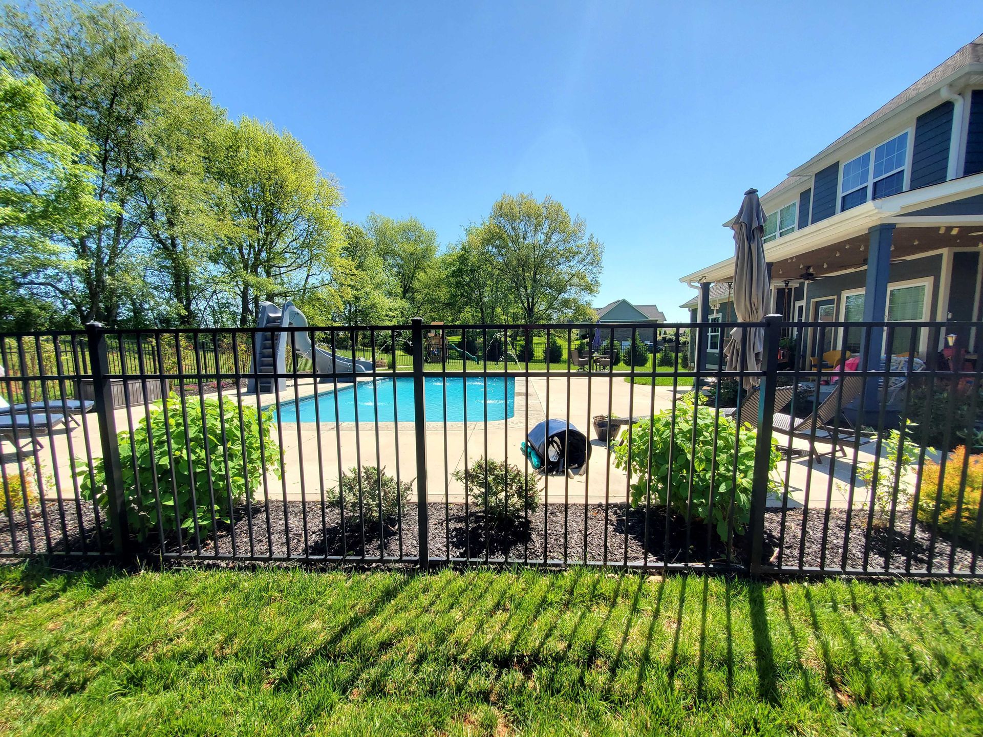 Black fence surrounds a backyard pool, lush greenery, and a house under a bright blue sky.