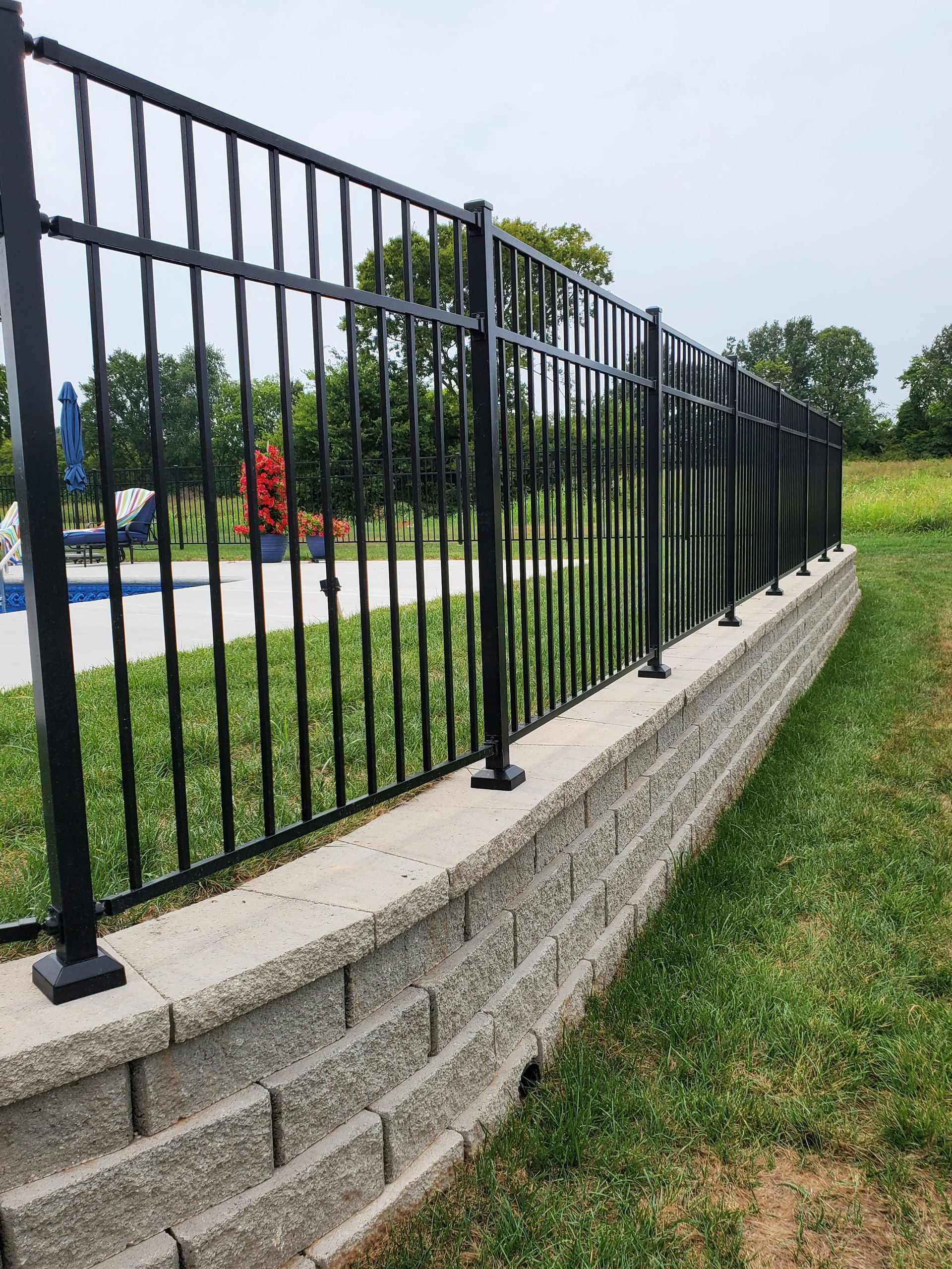 Black metal fence atop a retaining wall, bordering a grassy area and a pool.