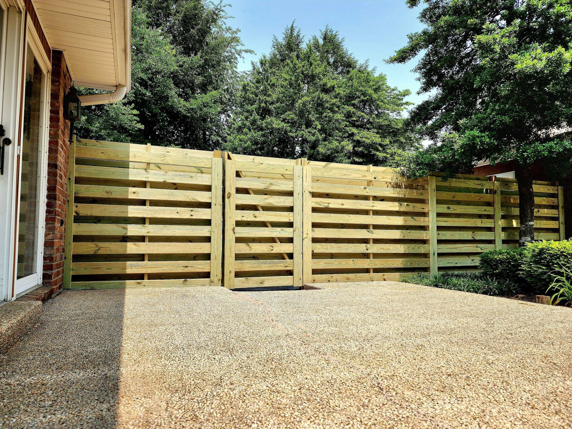 Gravel patio with a new wooden horizontal slat fence next to a brick wall, trees in the background.