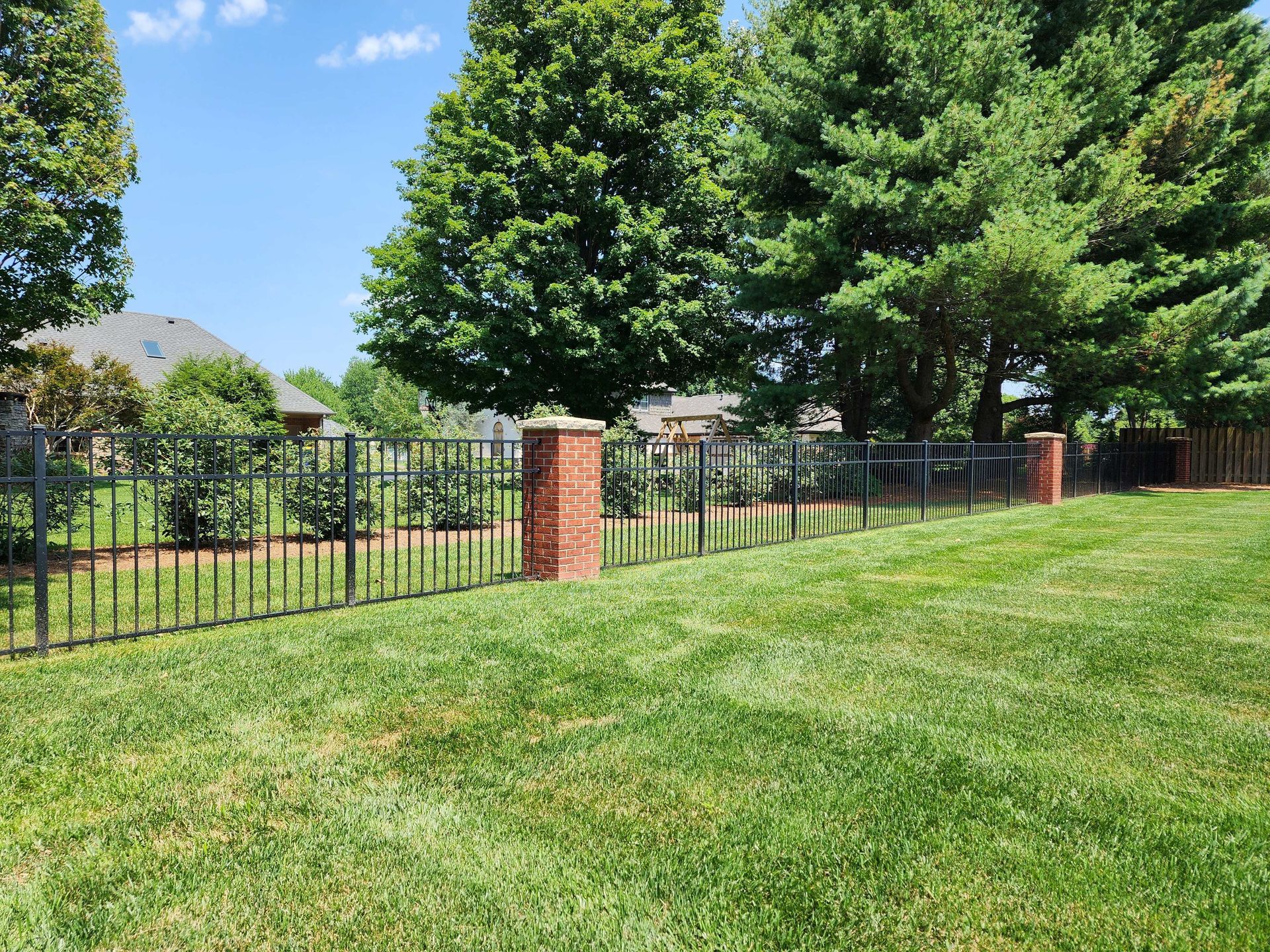 Lawn with black metal fence, brick pillars, and trees. A house is visible in the background on a sunny day.