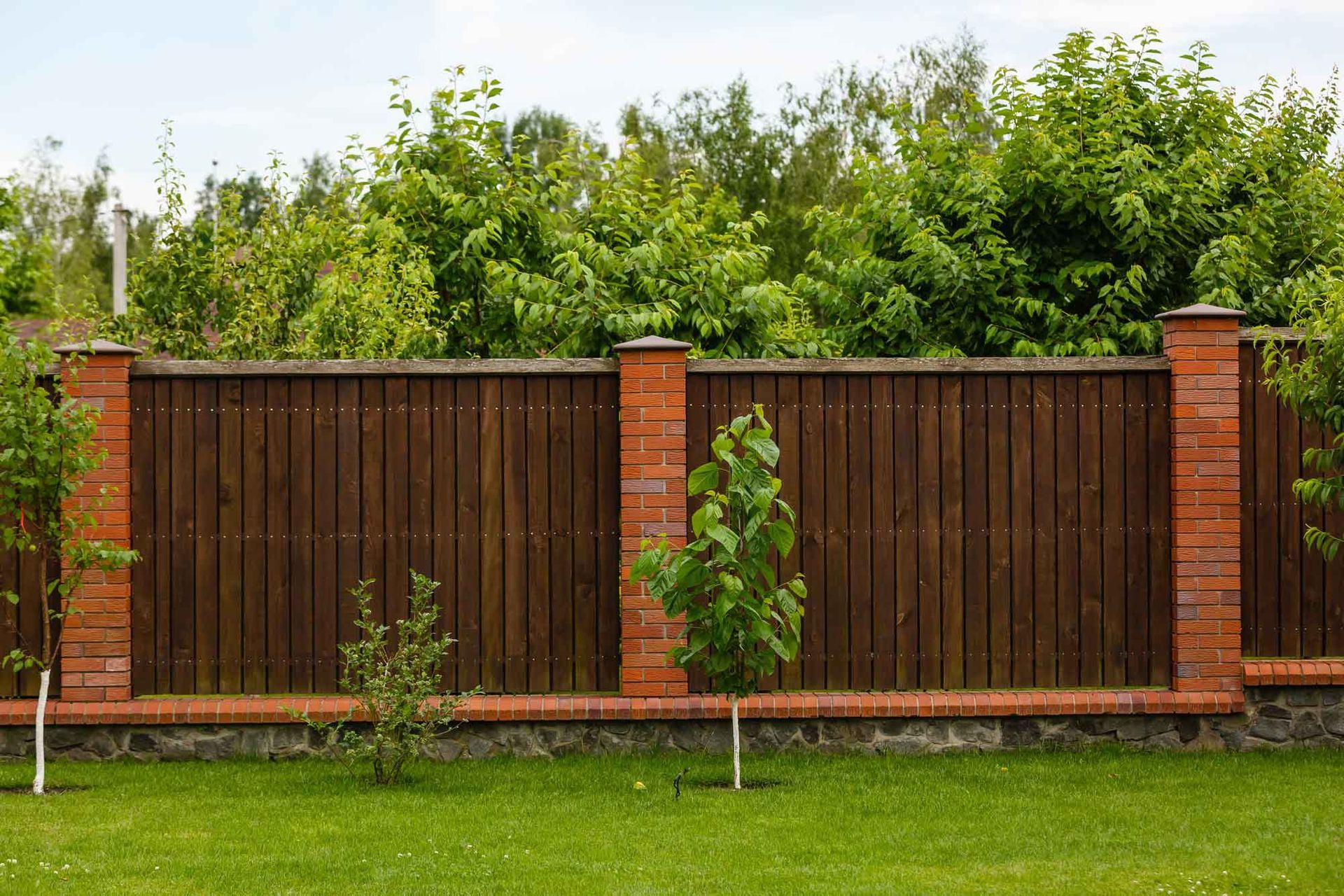 Wooden fence with brick pillars borders a green lawn and leafy garden.