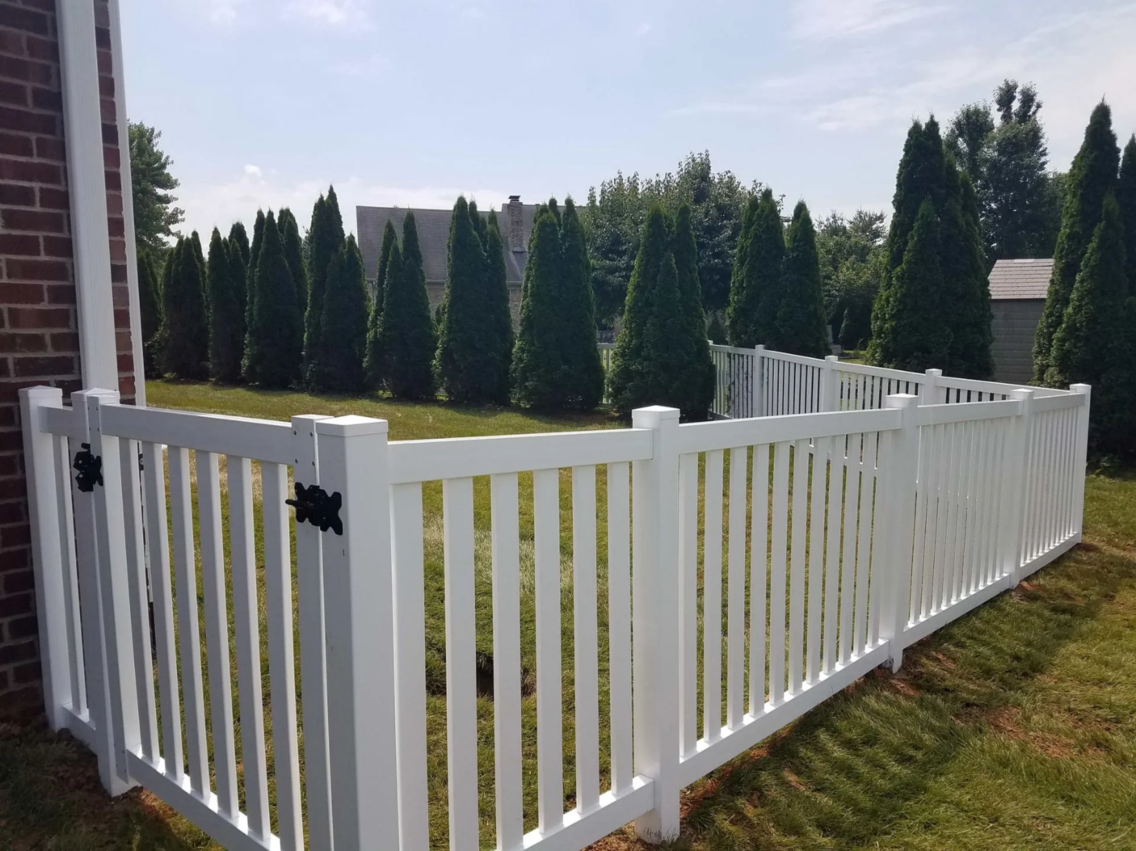 White picket fence surrounding a grassy yard with evergreen trees in the background.