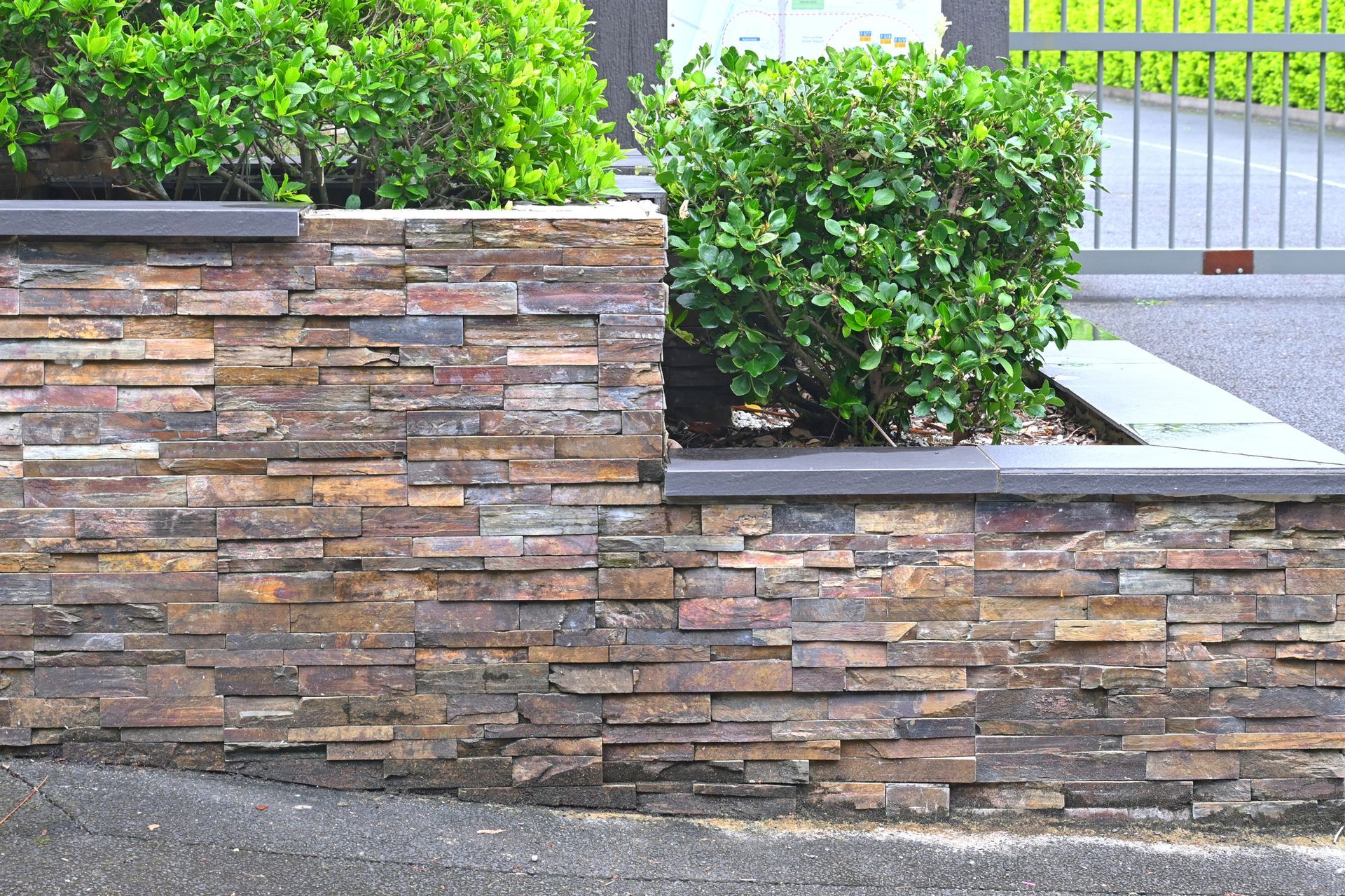 Stone retaining wall with green bushes in landscaped garden near gated entrance, outdoor design.