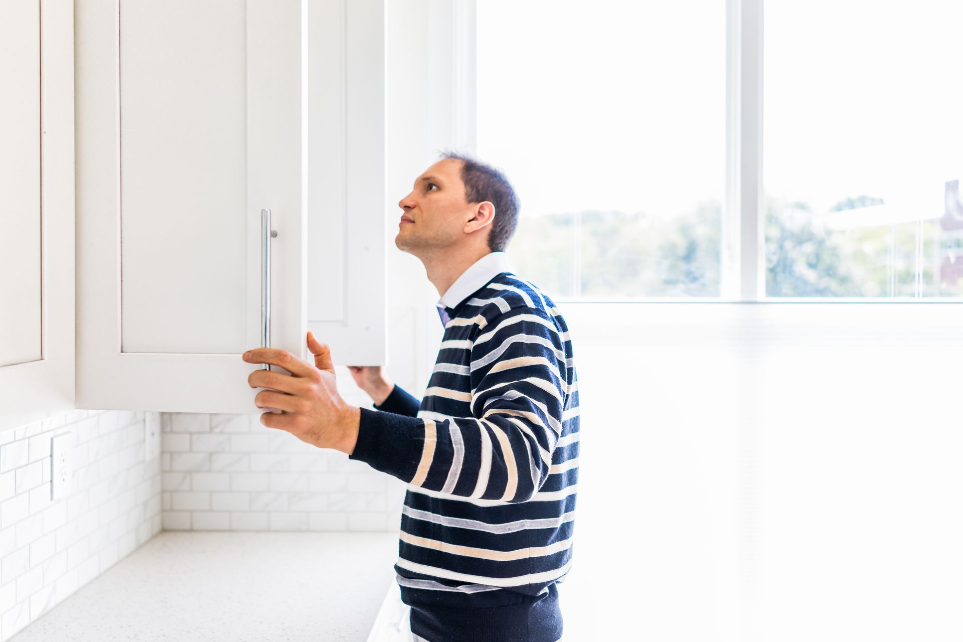 A man in a striped sweater is looking into a kitchen cabinet.