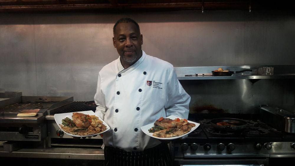Chef in a white jacket holding two plated dishes in a commercial kitchen.